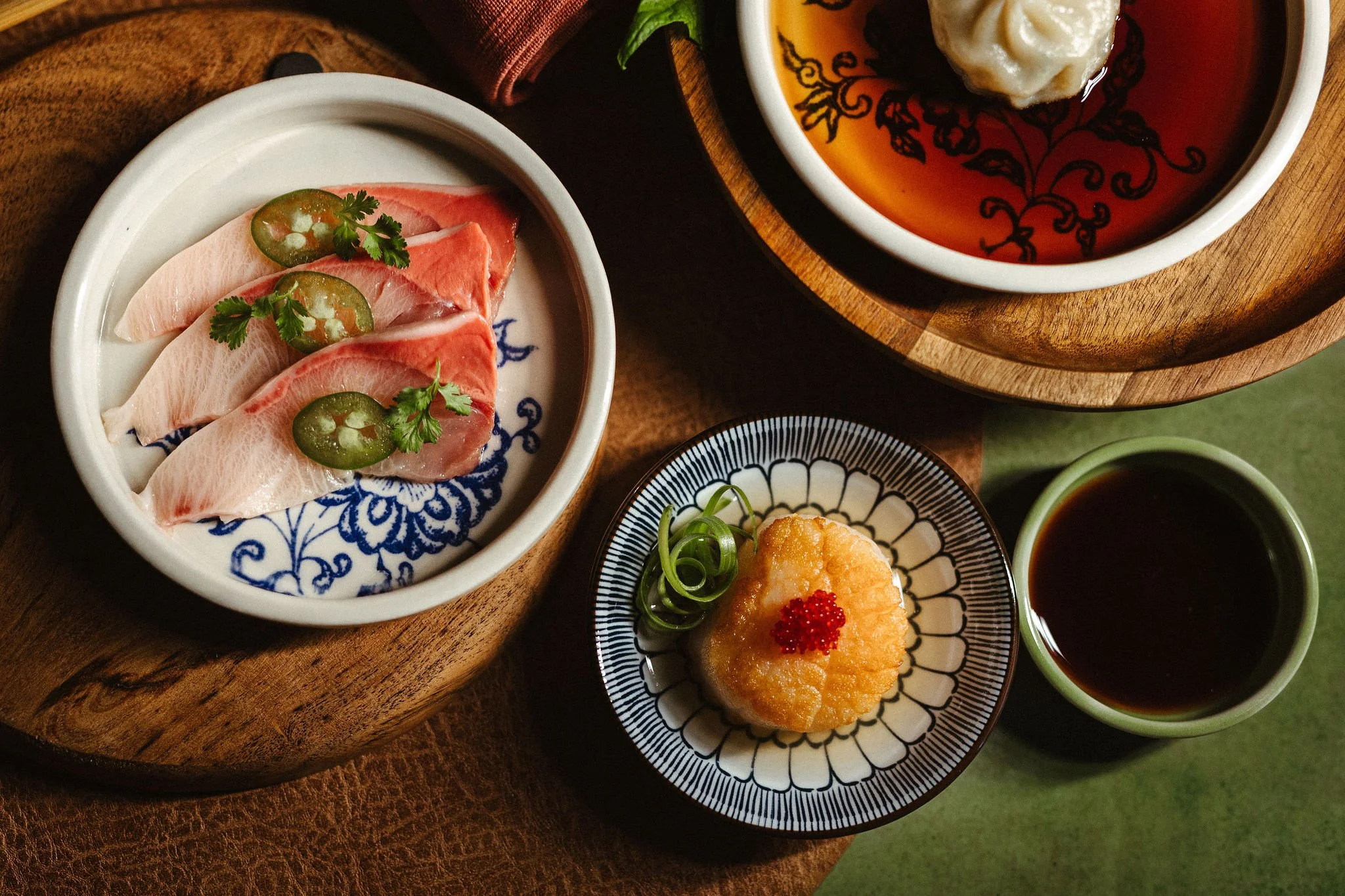 An assortment of Japanese dishes including sashimi garnished with jalapeño slices and herbs, a bowl of tempura topped with fish roe, a bowl of soup with dumplings, and small cups of dipping sauces on a wooden serving tray.