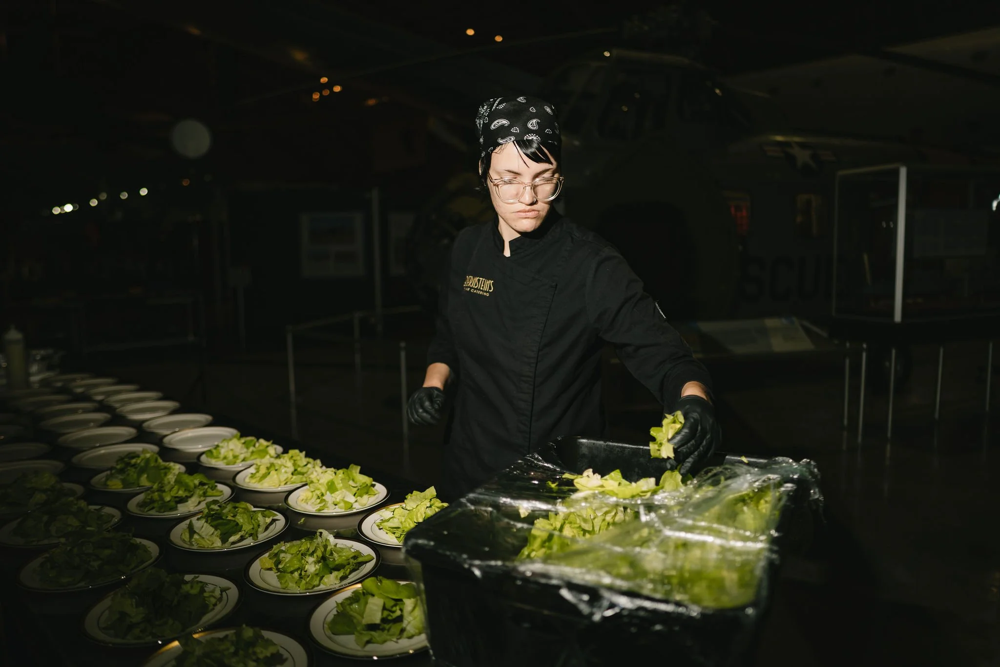 A woman wearing glasses, a black bandana, and black gloves prepares salads with lettuce and other greens at a dimly lit catering event or kitchen.