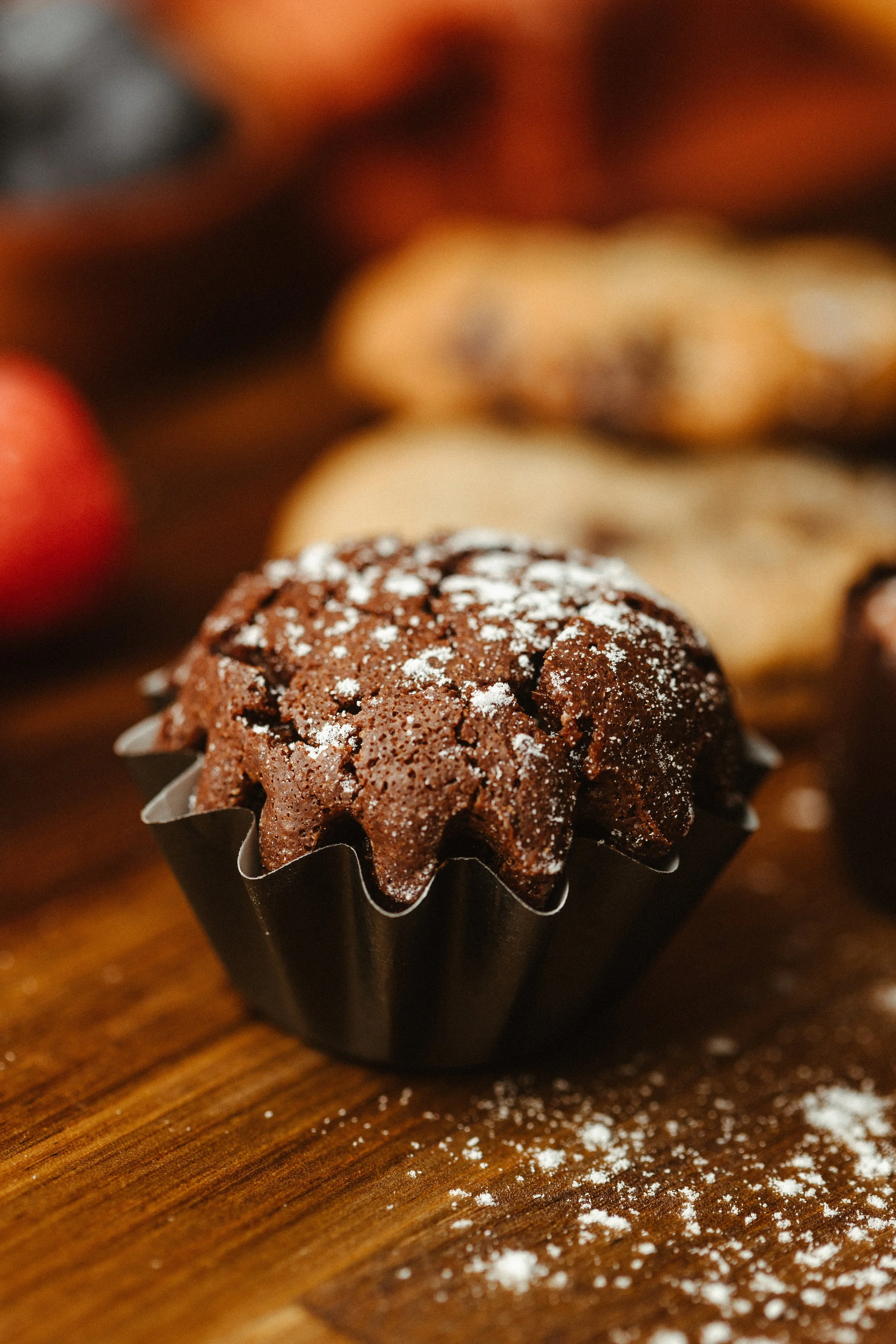 Close-up of a chocolate muffin topped with powdered sugar, placed in a black paper cup on a wooden surface with scattered powdered sugar.