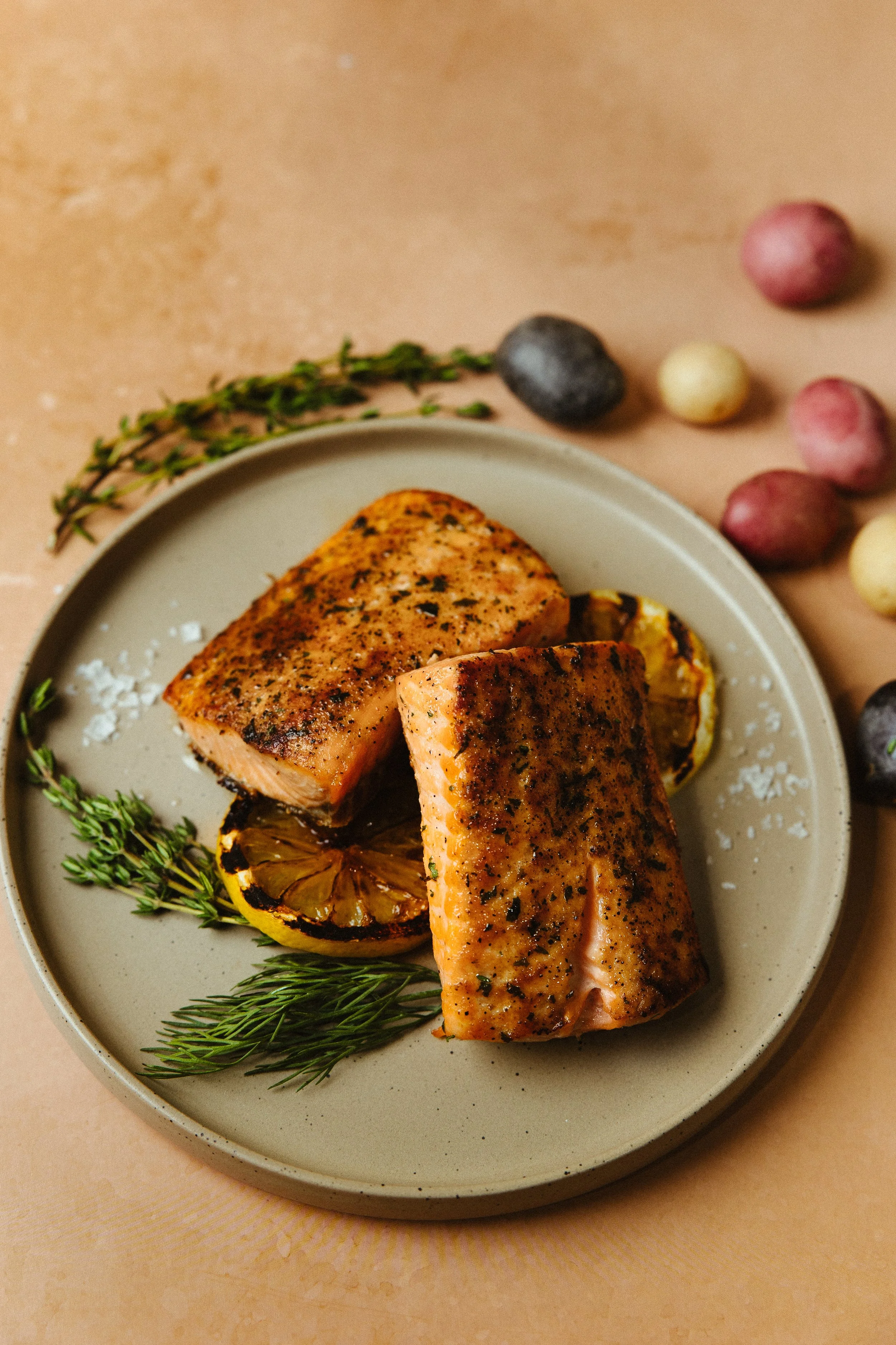 A plate with two pieces of cooked salmon seasoned with herbs, garnished with slices of grilled lemon and sprigs of fresh rosemary, on a beige surface with colorful small round potatoes and a sprig of thyme in the background.
