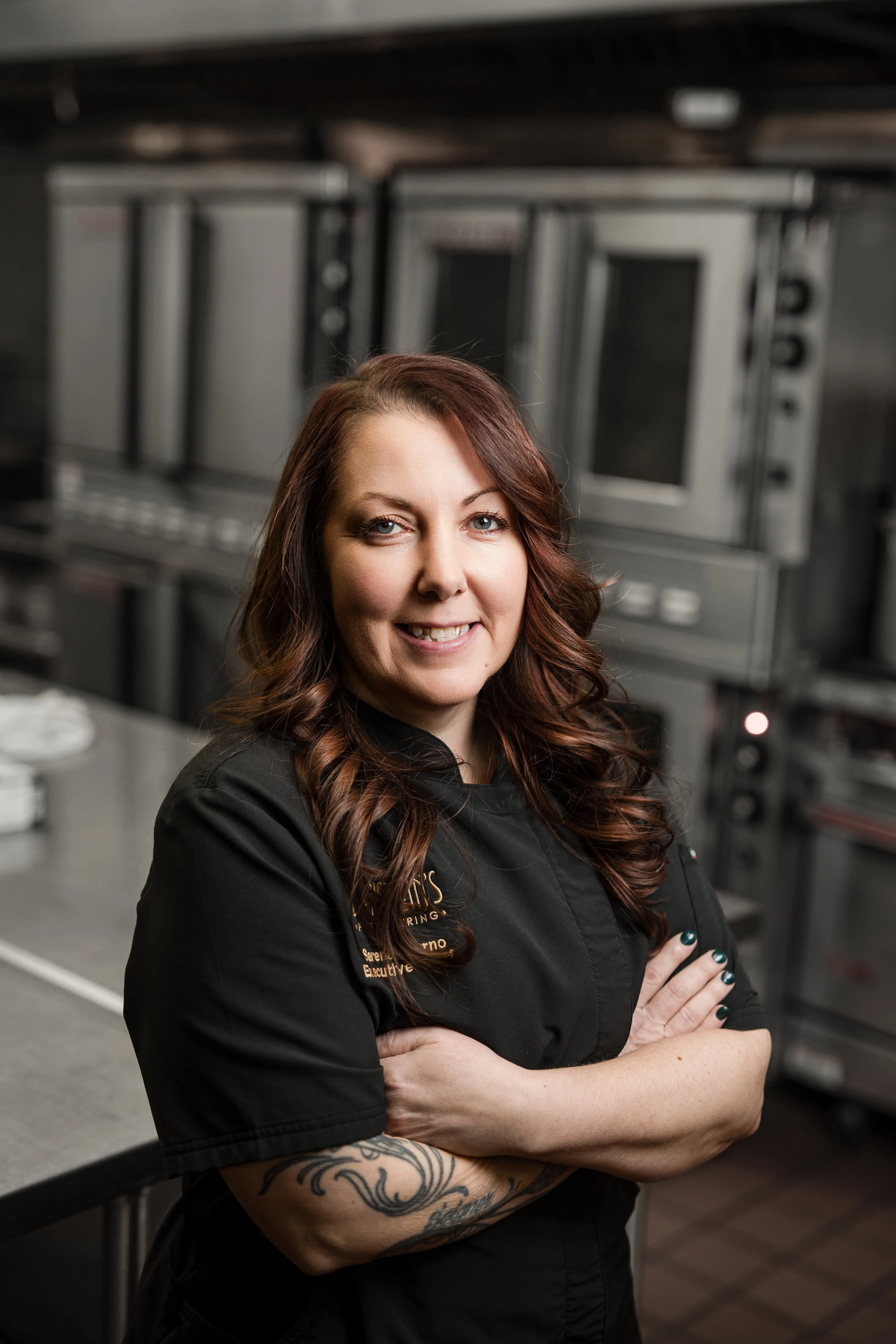 A woman with long wavy brown hair wearing a black chef's coat, standing with arms crossed in a professional kitchen.