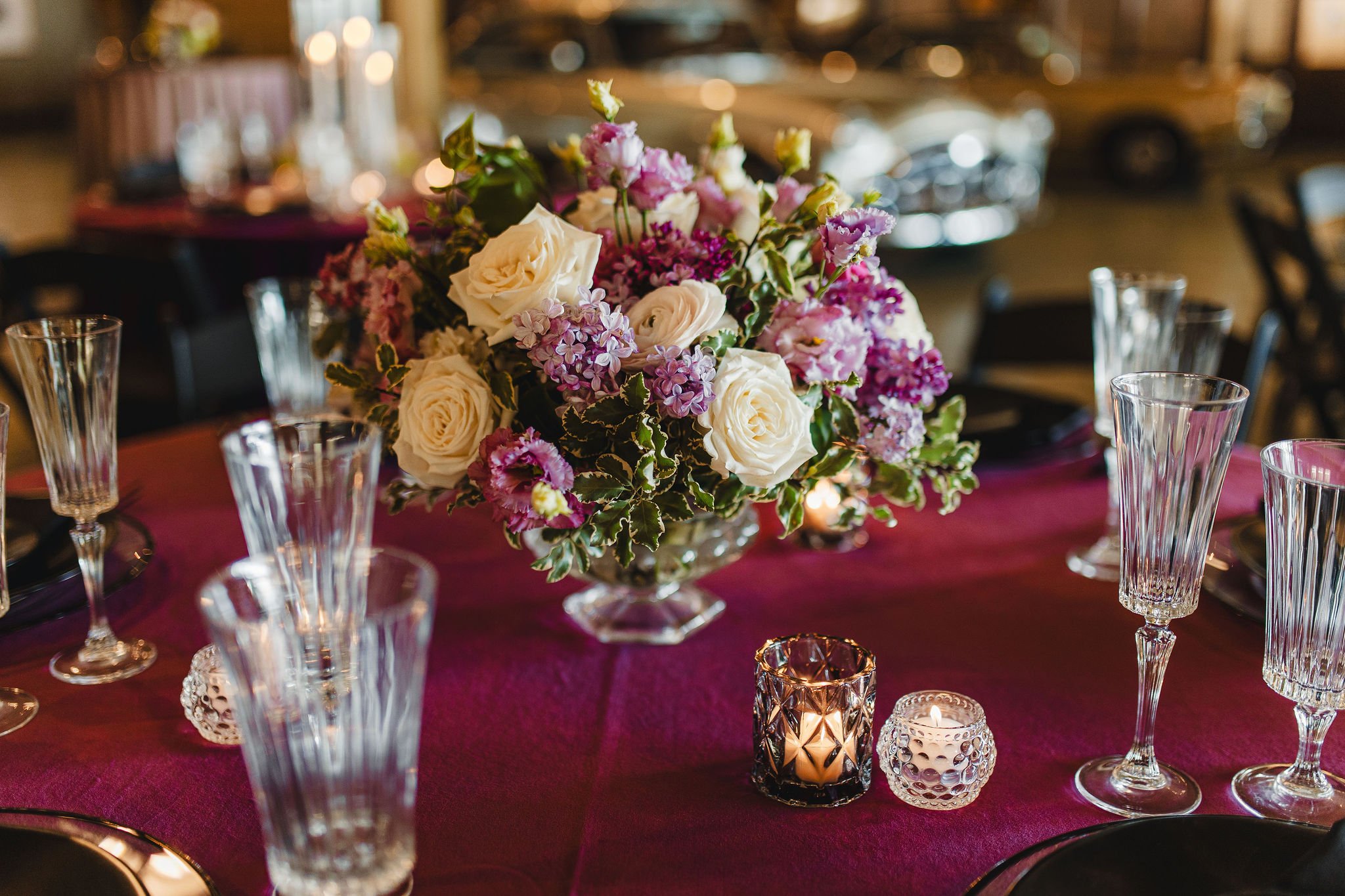 A floral centerpiece on a dining table with pink, white, and purple flowers, surrounding glassware and candles in a dimly lit room.
