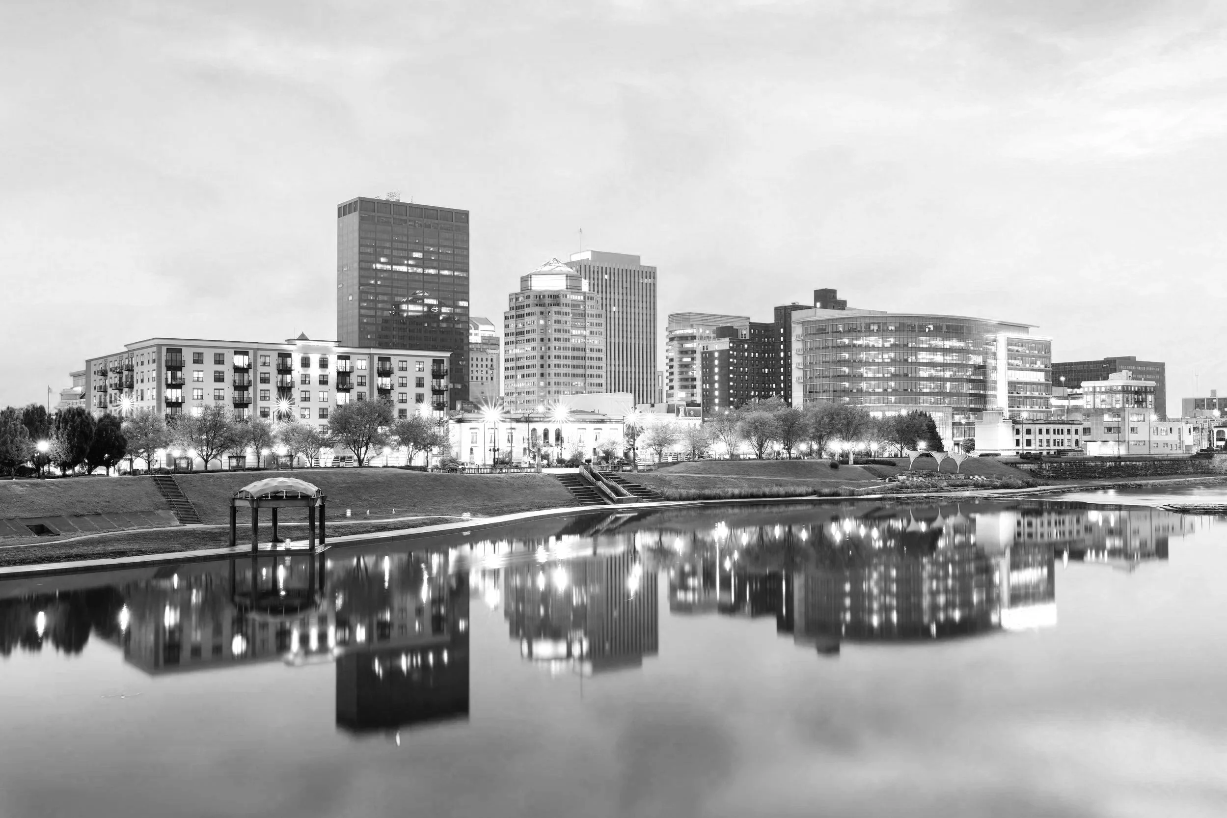 Black and white photograph of a city skyline with tall modern buildings viewed from across a calm river. The buildings' reflections are visible in the water, and a park with trees and benches is in the foreground.
