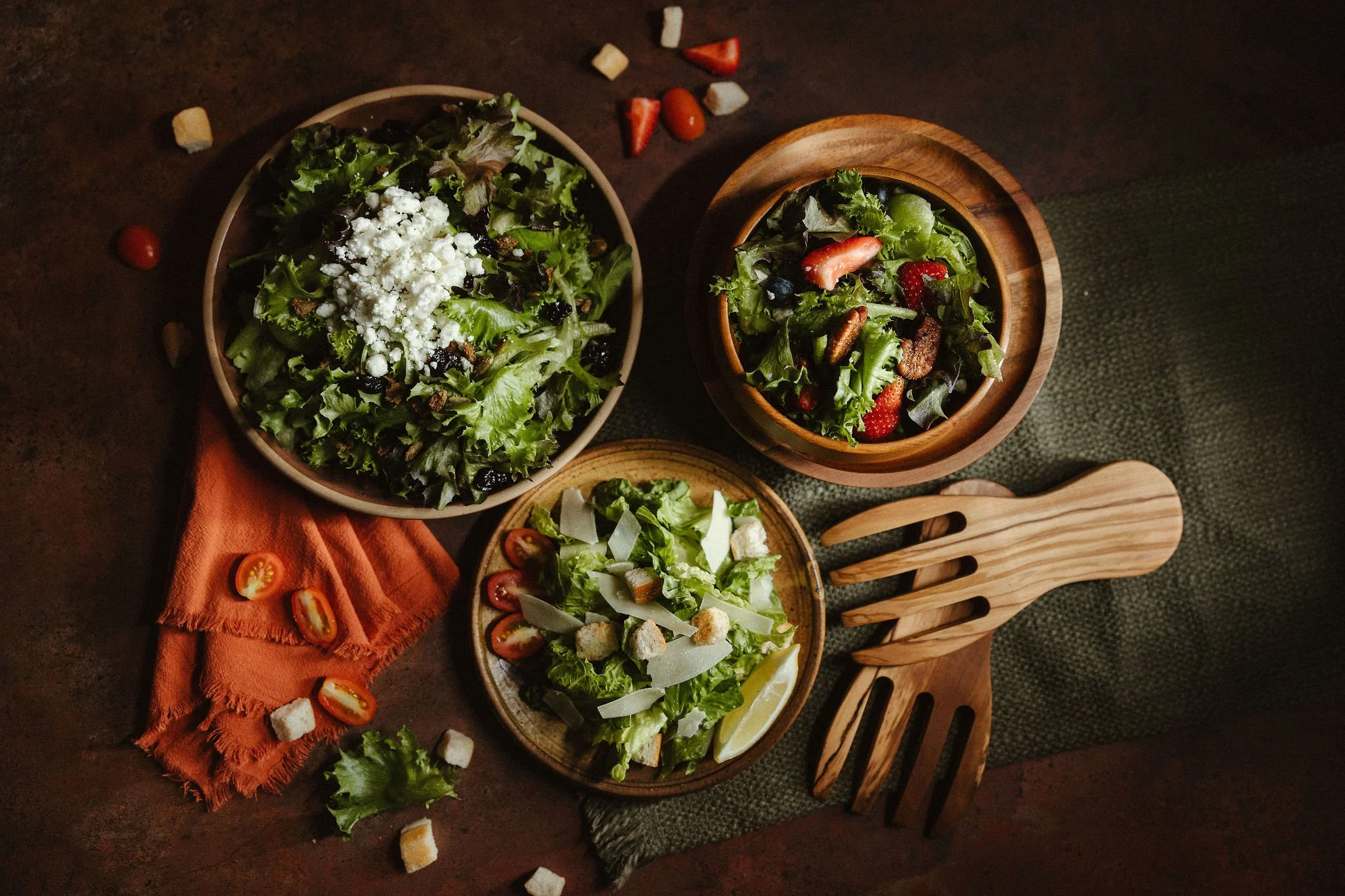 Several bowls of fresh salad on a dark wooden table, with grape tomatoes and croutons scattered around. One bowl contains leafy greens, strawberries, blueberries, and bacon bits, while others have assorted salad ingredients with cheese and dressing.