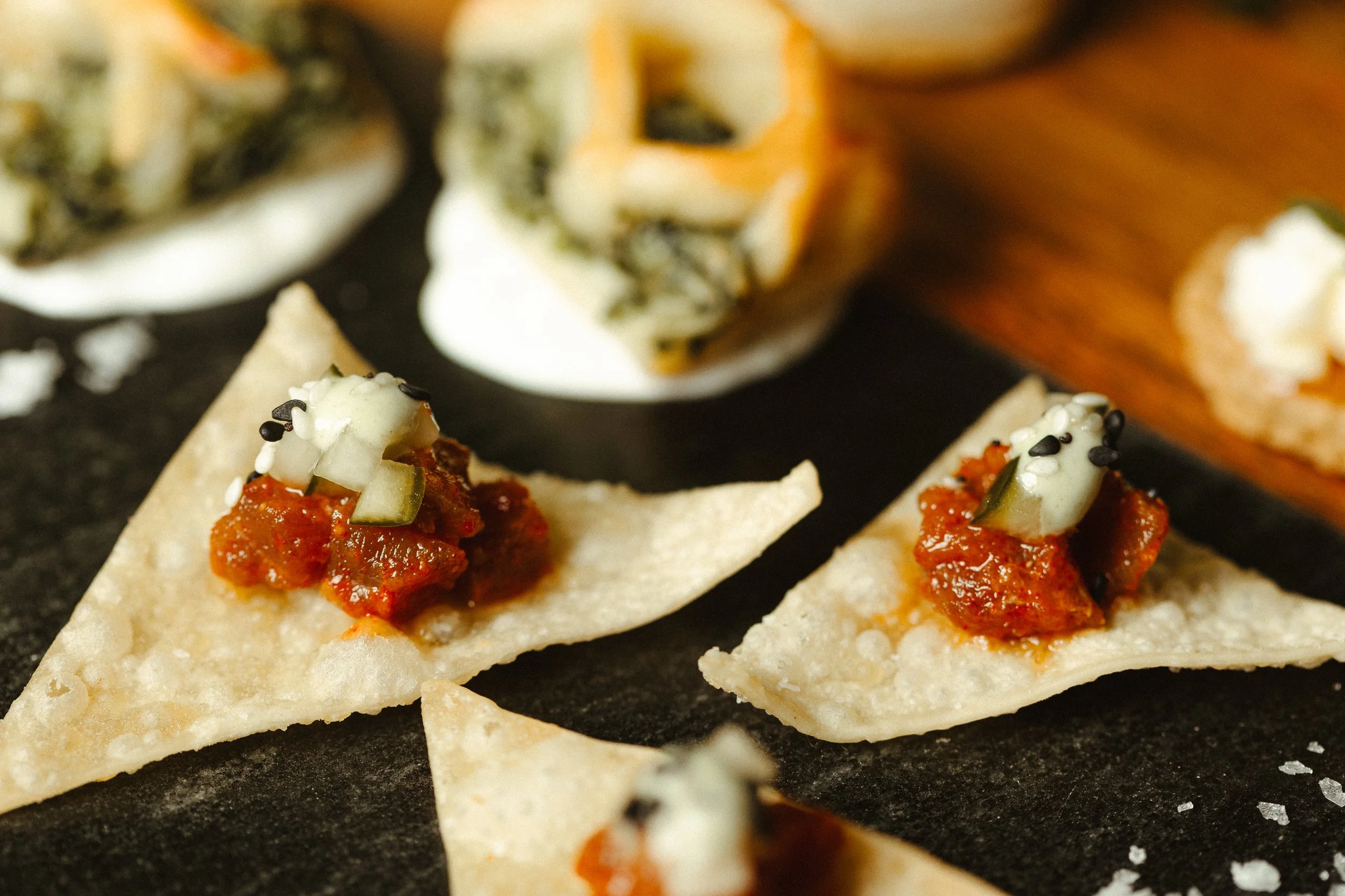 Close-up of bite-sized tortilla chips topped with red salsa, chopped onions, and garnish, on a dark surface.