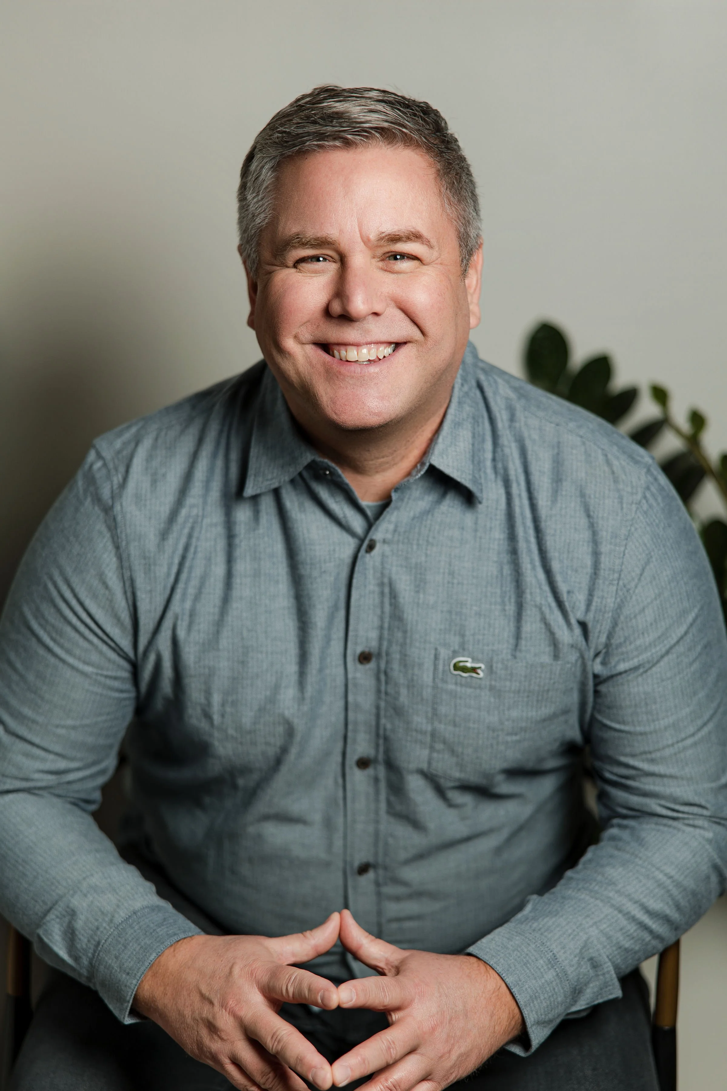 A smiling man in a gray shirt sitting with his hands clasped on his lap.