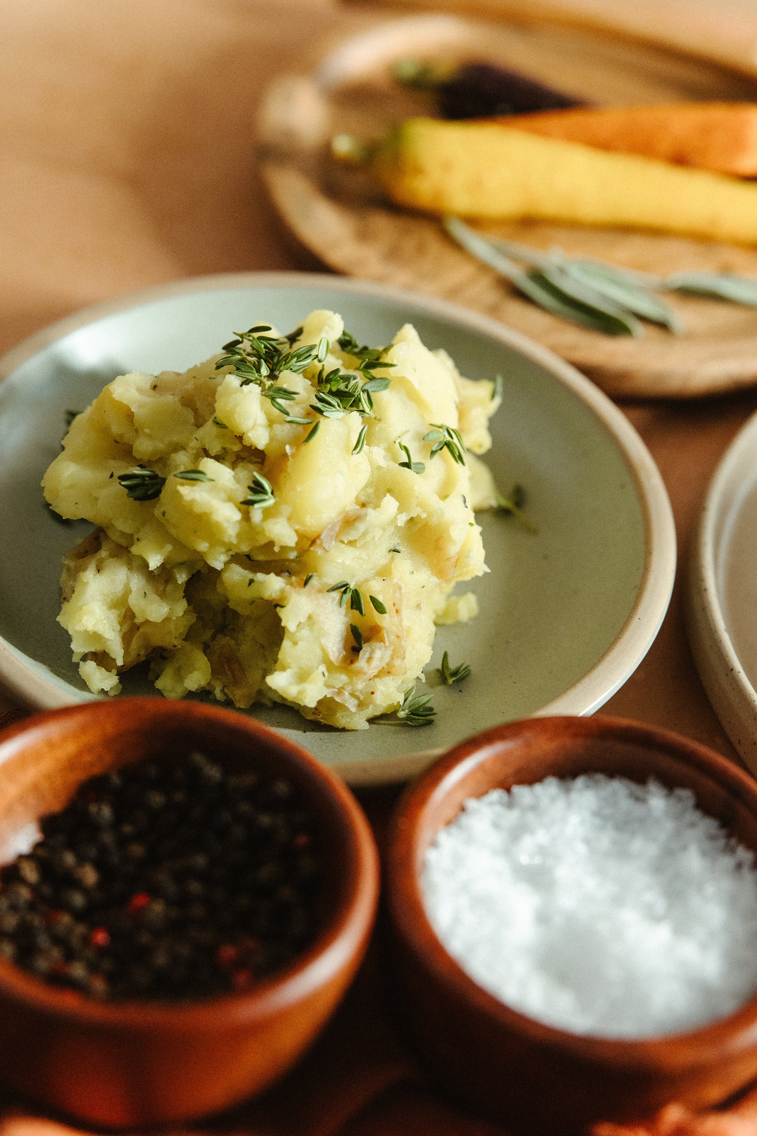 A bowl of mashed potatoes garnished with fresh herbs, with a wooden platter of sliced vegetables and bowls of black pepper and salt in the background.