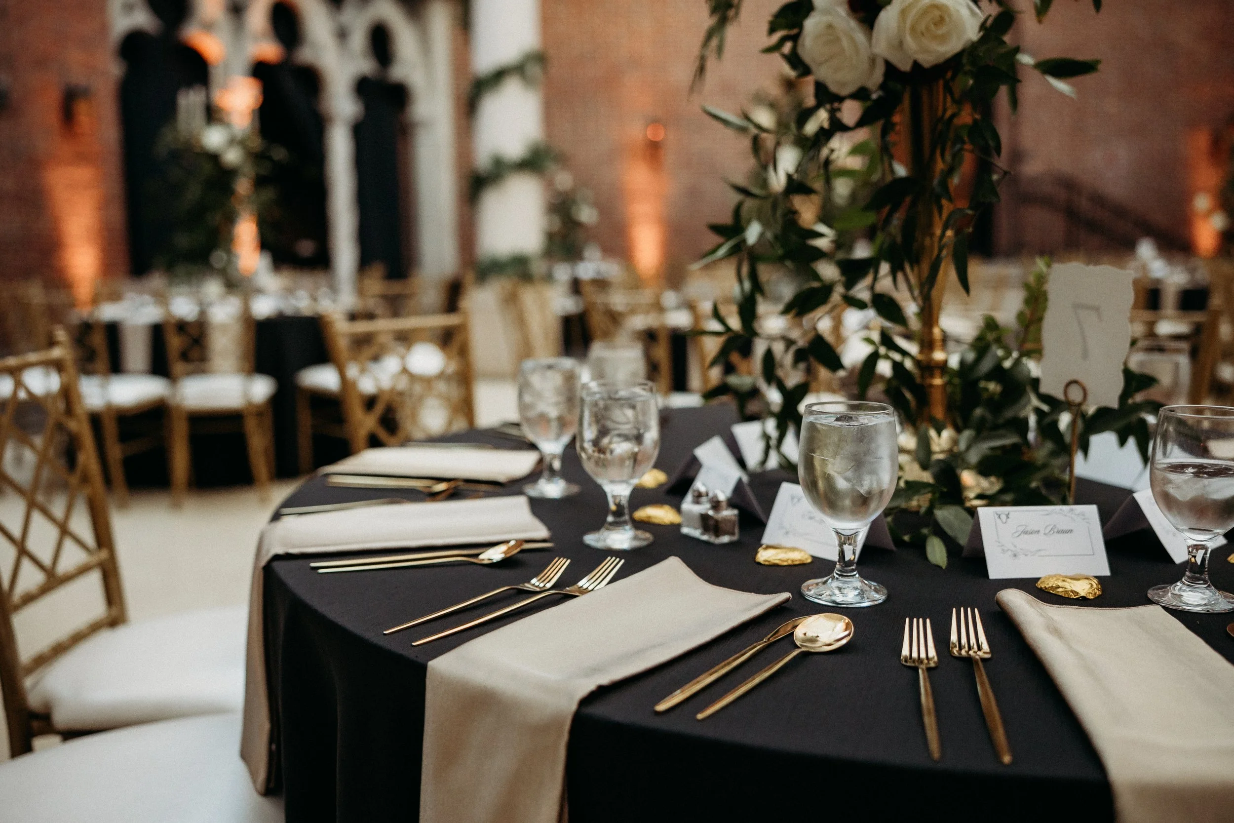 Elegant banquet table with black and cream table settings, gold cutlery, glassware, and floral centerpieces in a decorated event space with chairs and warm lighting.