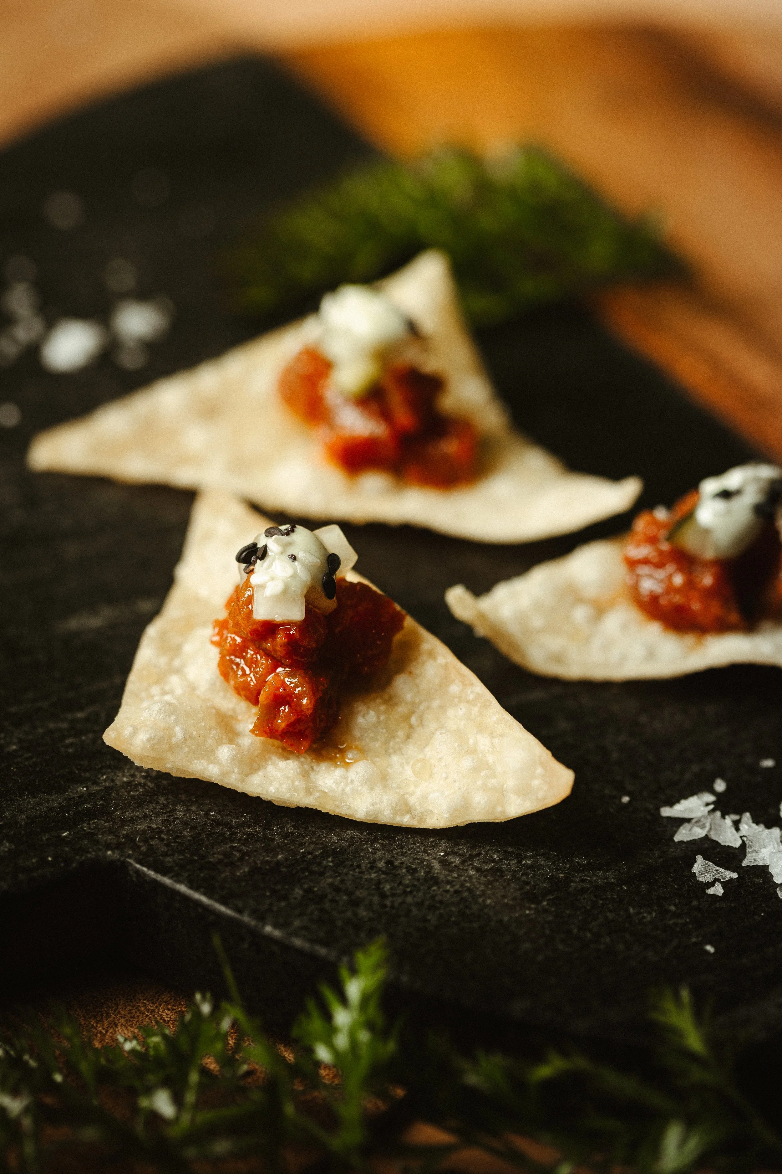 Close-up of tortilla chips topped with salsa, sour cream, and black sesame seeds on a black serving plate, surrounded by green pine branches.