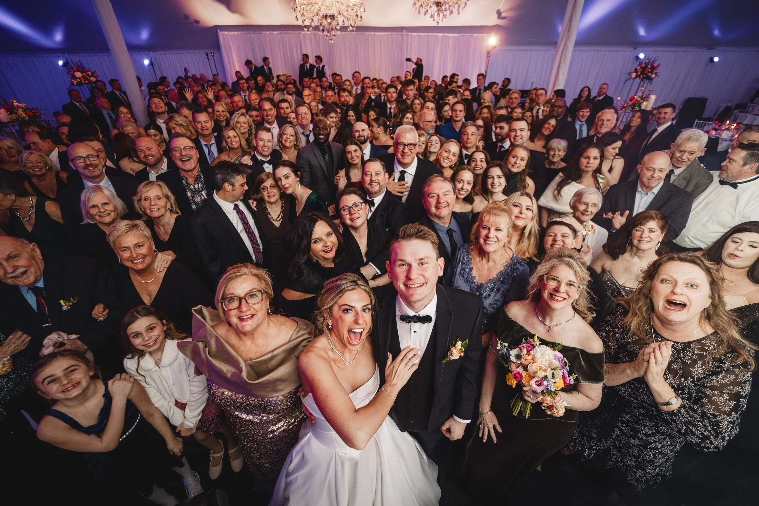 A large group of people dressed in formal attire at a celebration or wedding, smiling and posing together in a decorated event hall with chandeliers and floral arrangements.
