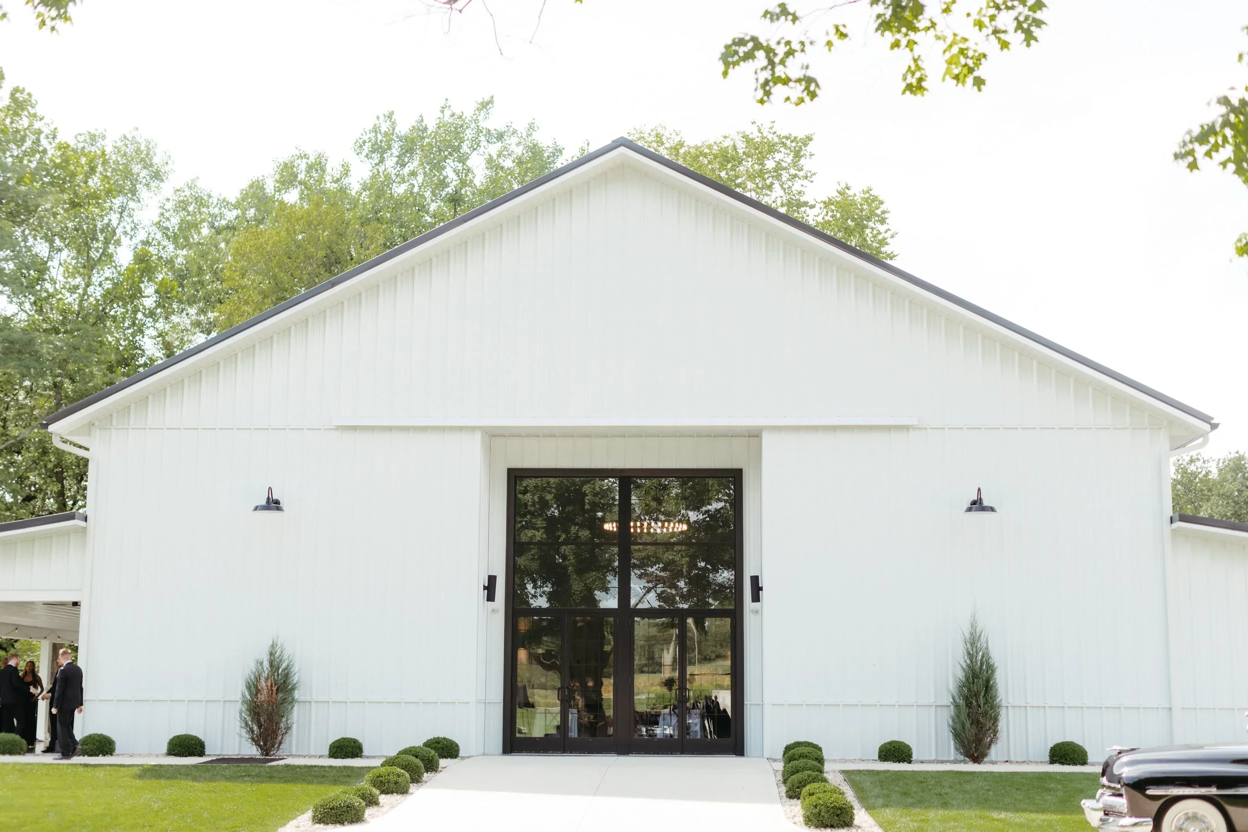 Front view of a modern white building with black framed glass doors, surrounded by a well-manicured lawn and small bushes, with trees in the background.