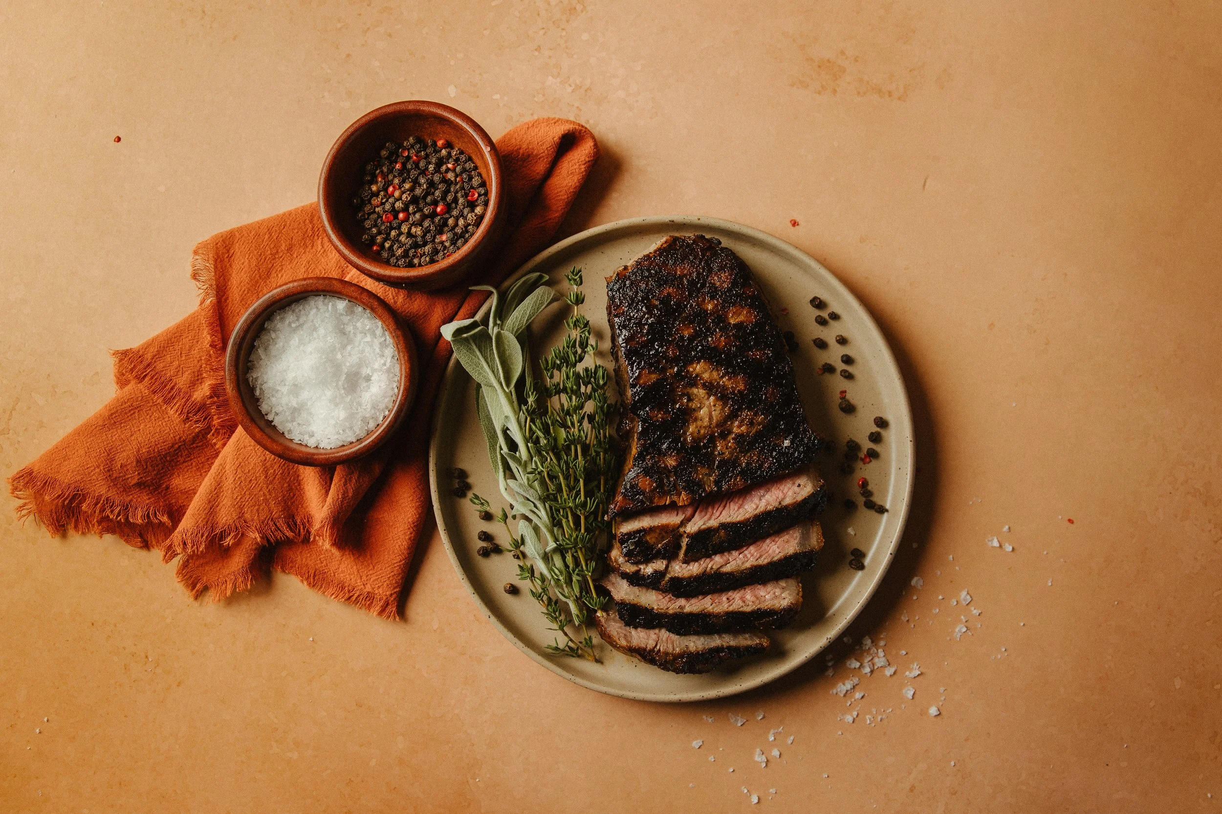Sliced grilled steak garnished with herbs on a white plate, accompanied by salt and pepper shakers and a sprig of herbs on a beige surface with an orange napkin.