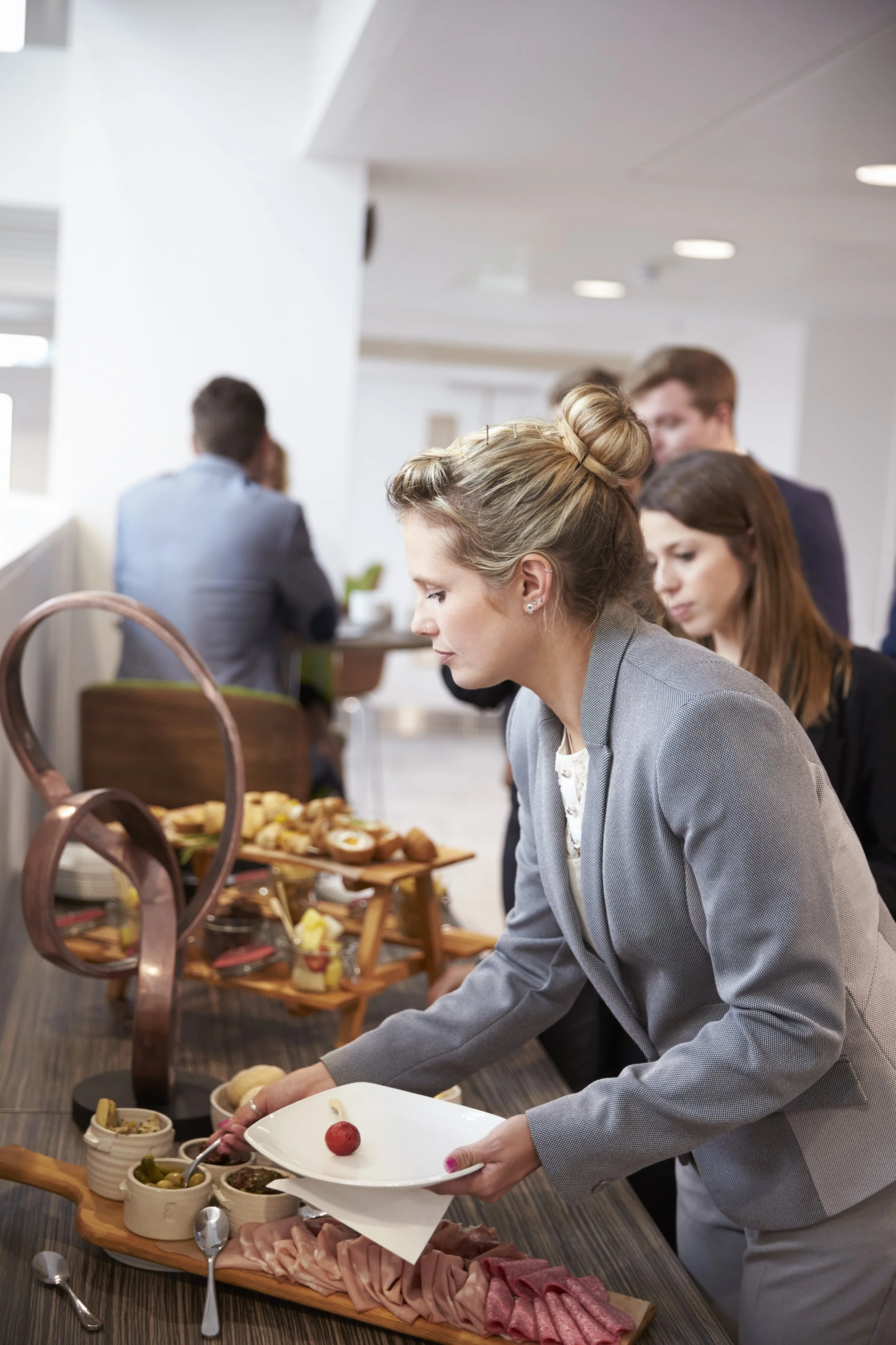 A woman in a gray blazer serving herself food from a buffet table with various dishes, including sliced meats and accompaniments, in a well-lit indoor setting.