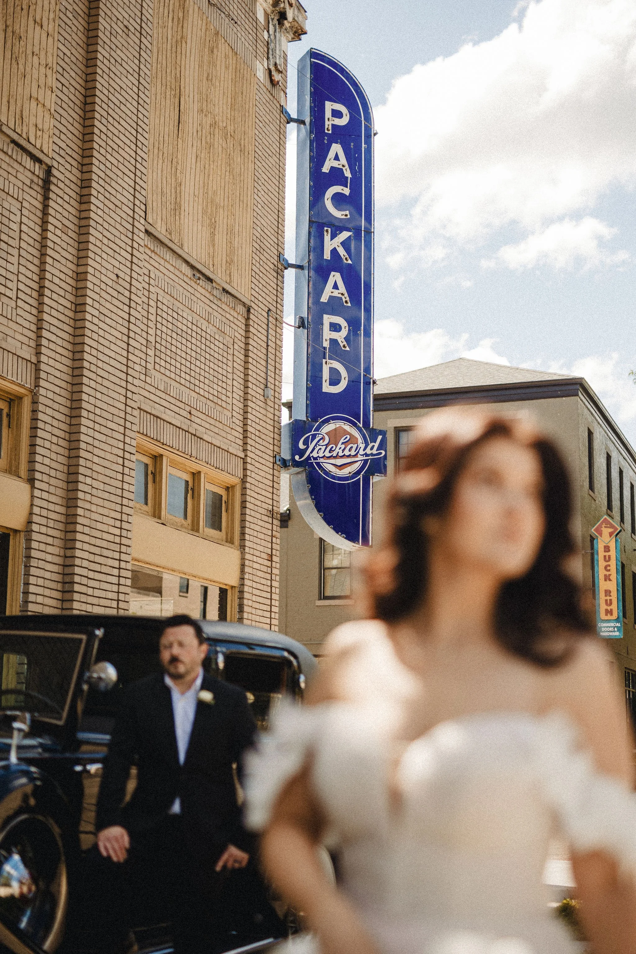 A woman in a wedding dress walking outdoors on a city street with buildings and a blue parking garage sign in the background.