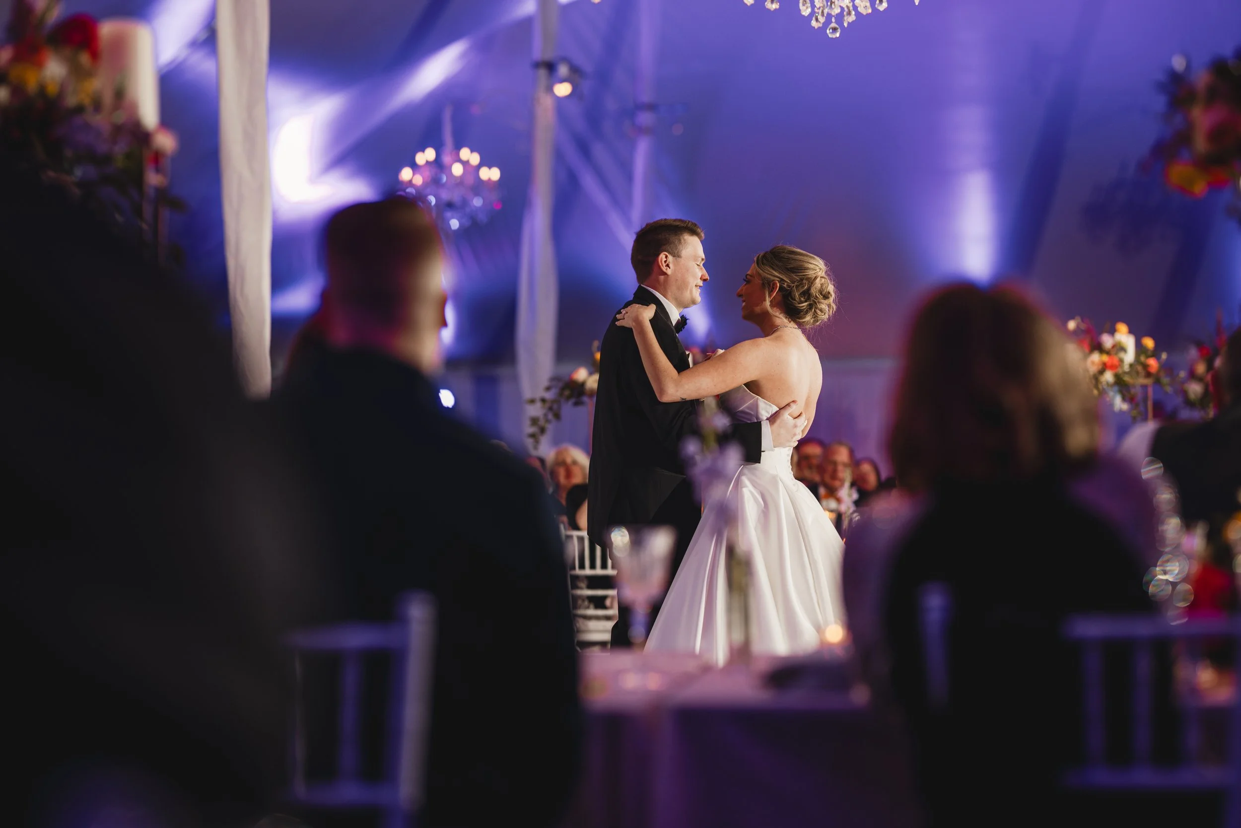 A bride and groom share a dance at their wedding reception inside a decorated tent with purple lighting. The bride wears a strapless wedding gown, and the groom wears a tuxedo. Guests are seated around them, watching the couple dance.