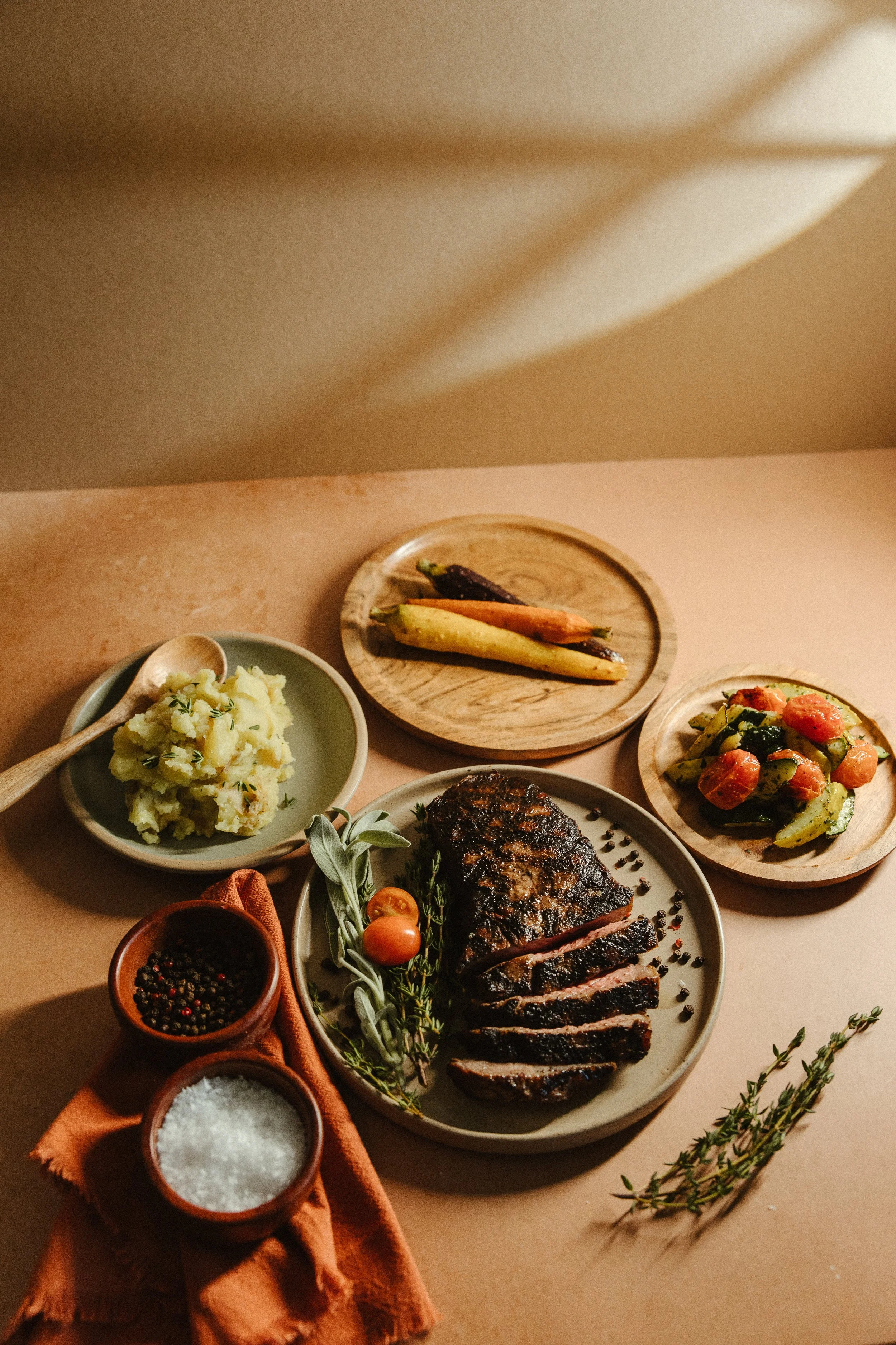 A table with various dishes including grilled meat, mashed potatoes, roasted vegetables, and condiments, with sprigs of herbs and cooking ingredients.
