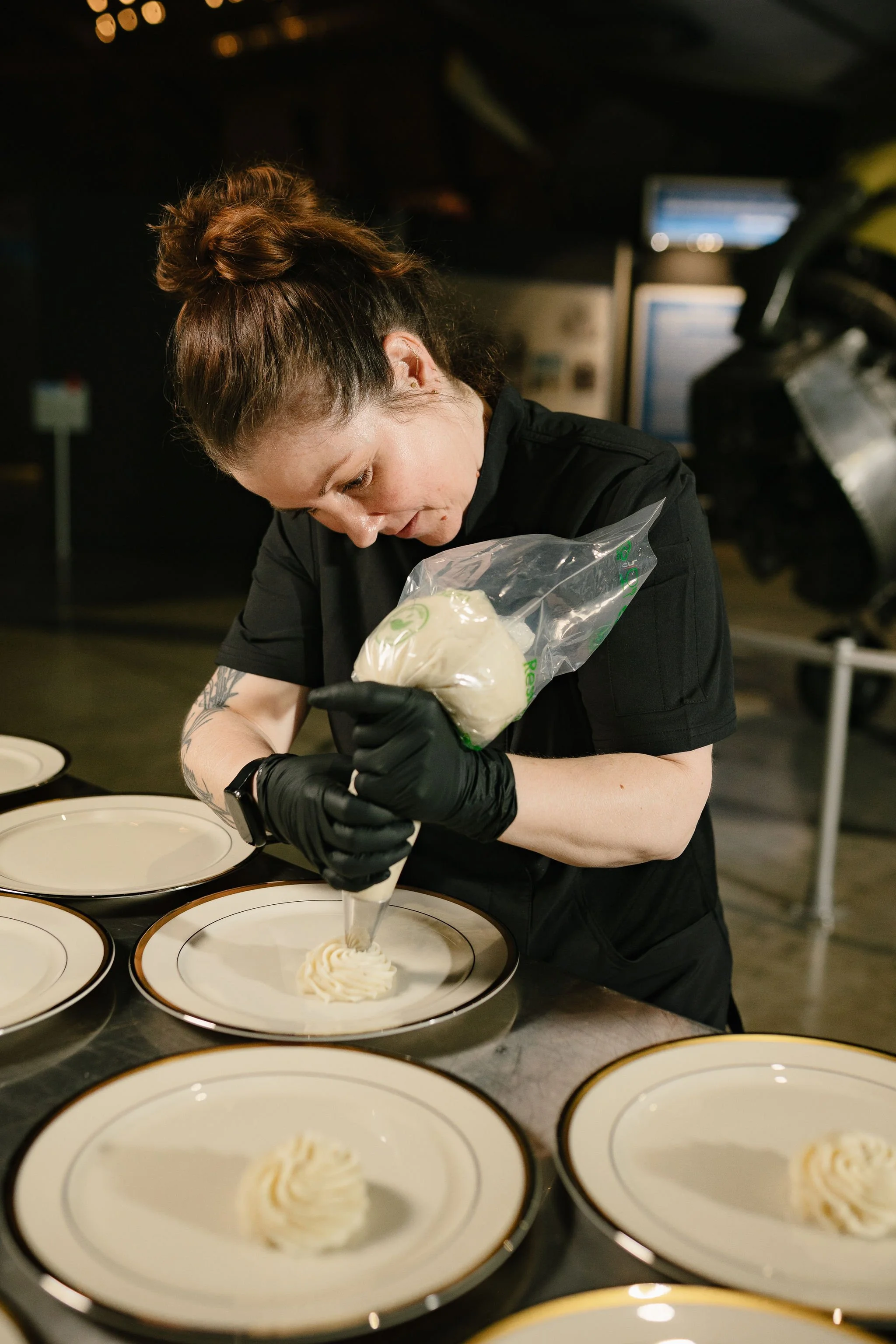 A woman with braided hair and tattoos on her arm is piping white frosting onto plates of dessert, wearing black gloves, in a professional kitchen setting with multiple similar plates prepared on a table.