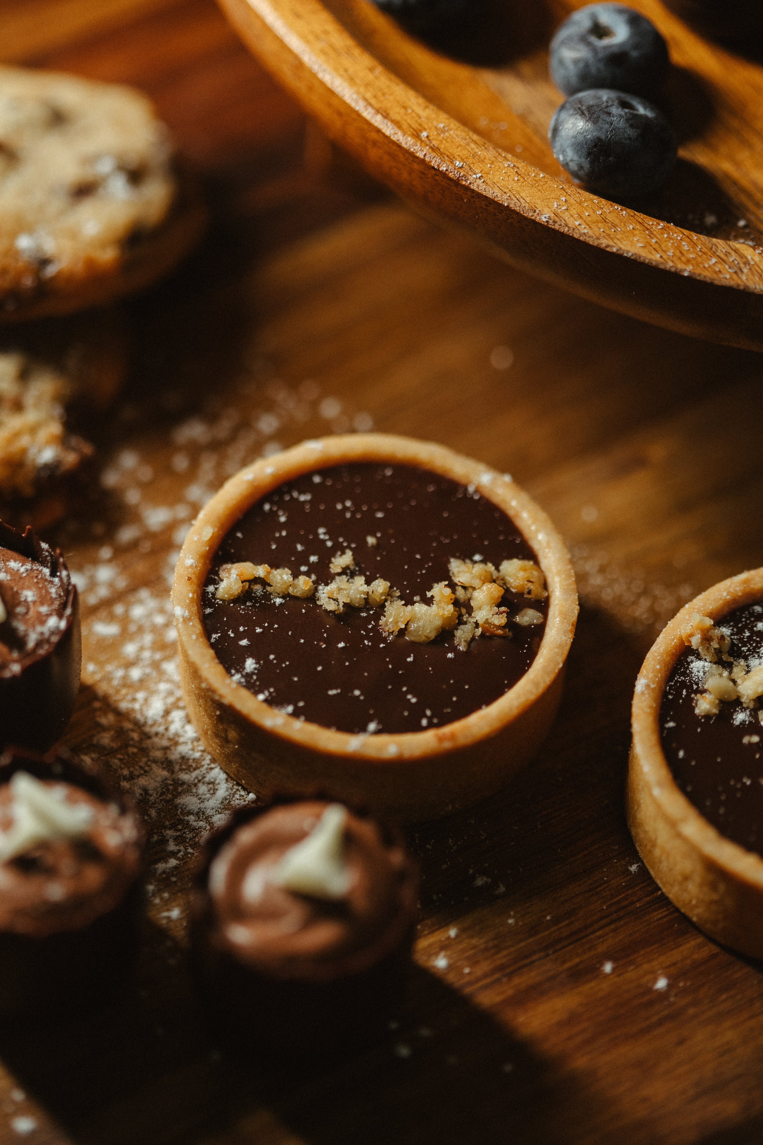 Close-up of small chocolate tartlets topped with chopped nuts on a wooden surface, with a few blueberries and a wooden bowl visible in the background.