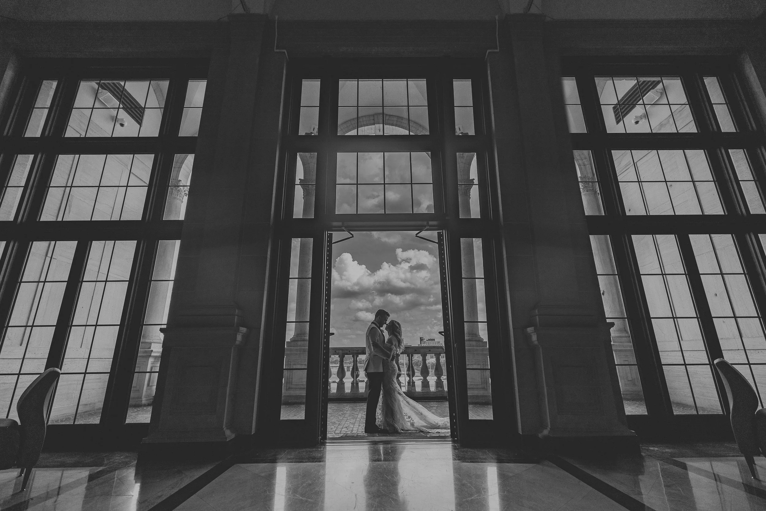 A couple dressed in wedding attire facing each other in front of a large window with a view outside, in a grand indoor setting with a high ceiling and arches.