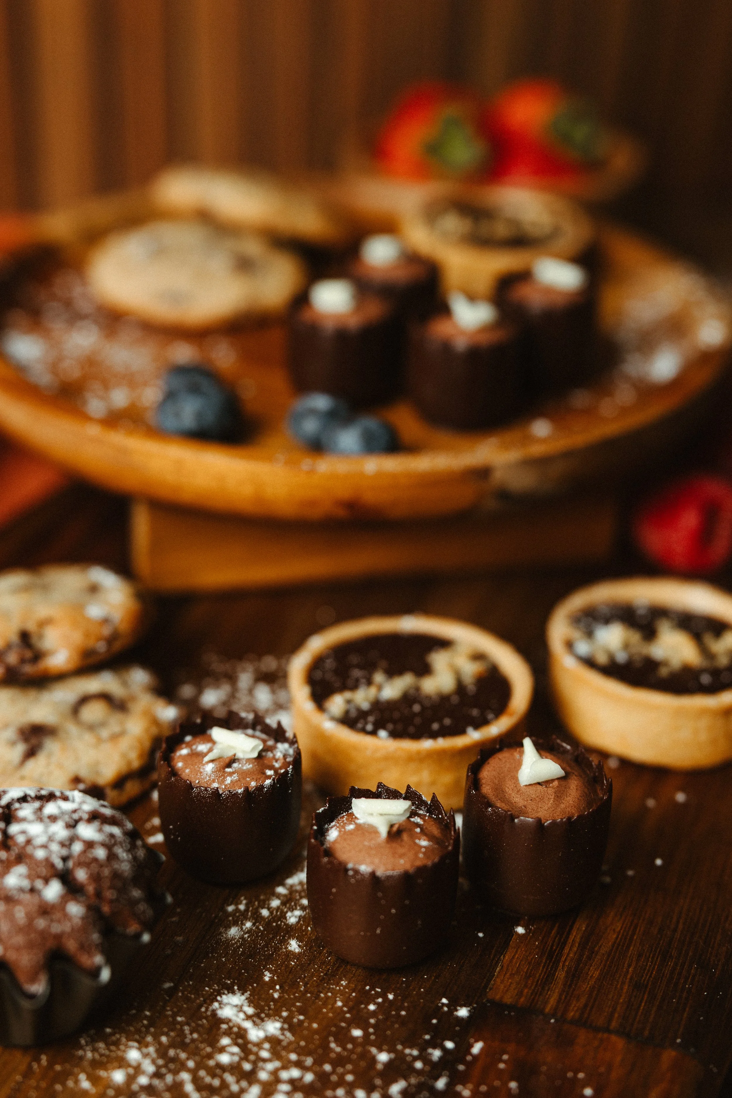 Assorted miniature desserts, including chocolate cups, tartlets, and cookies, displayed on a wooden platter with strawberries in the background.