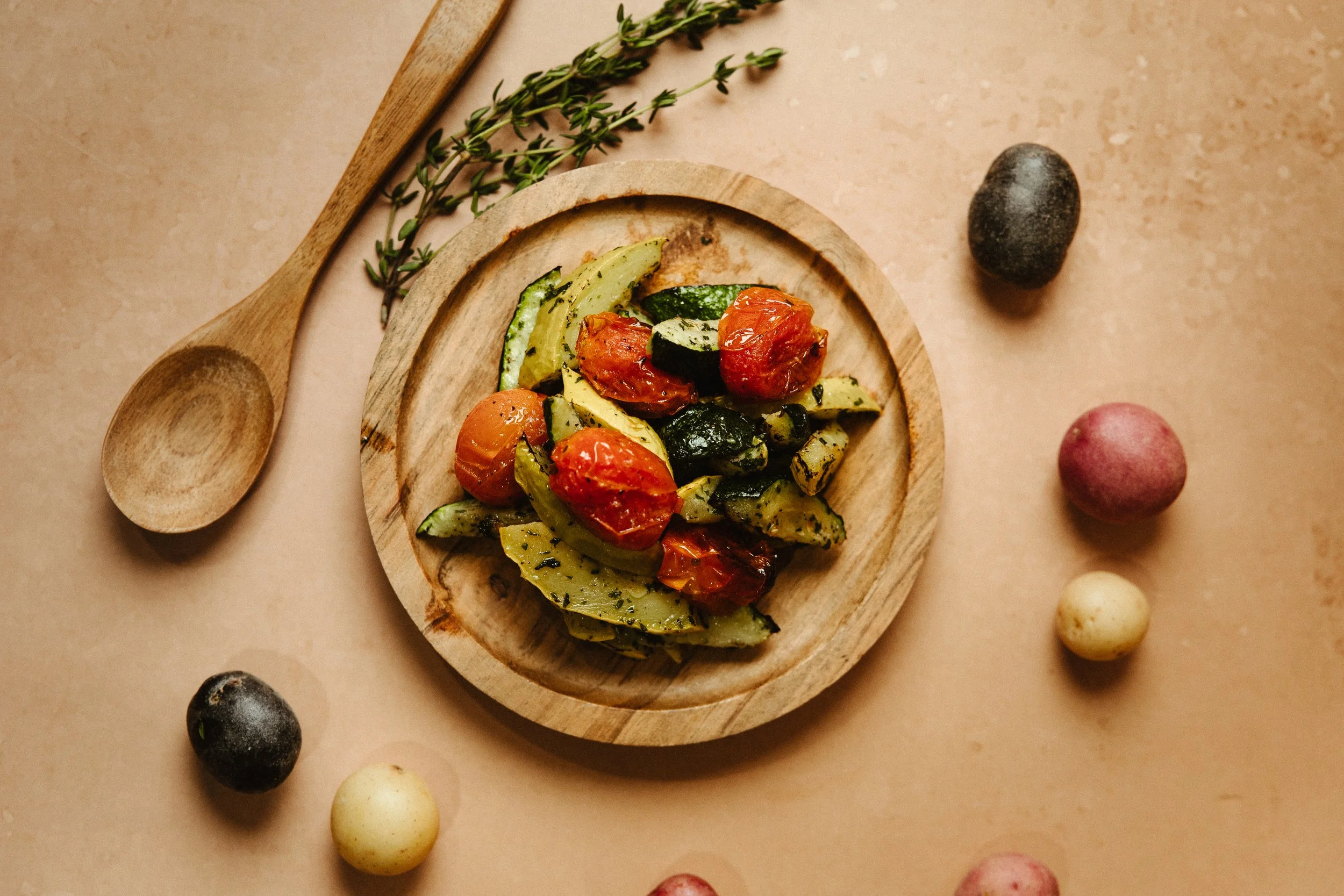 A wooden plate with grilled vegetables including cherry tomatoes, zucchini slices, and zucchini chunks, garnished with herbs. Surrounding the plate are scattered colorful stones and a sprig of thyme on a beige surface.