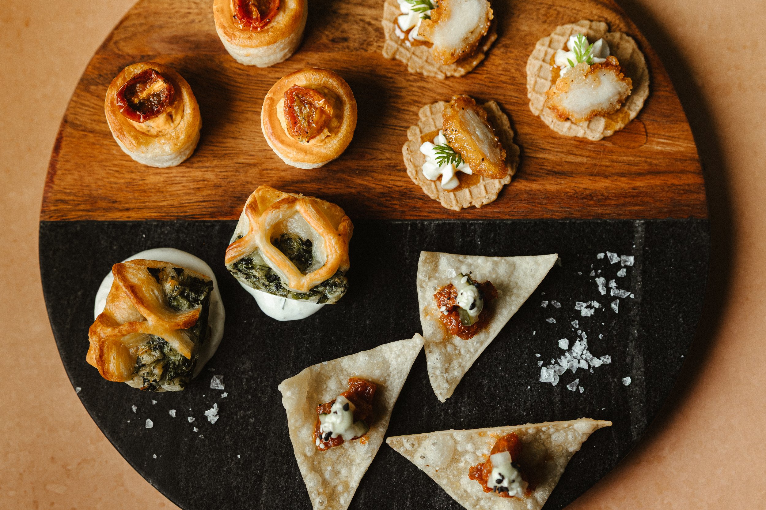 Assorted appetizers on a wooden and black slate serving platter, including mini pies, tartlets, and crackers with toppings.