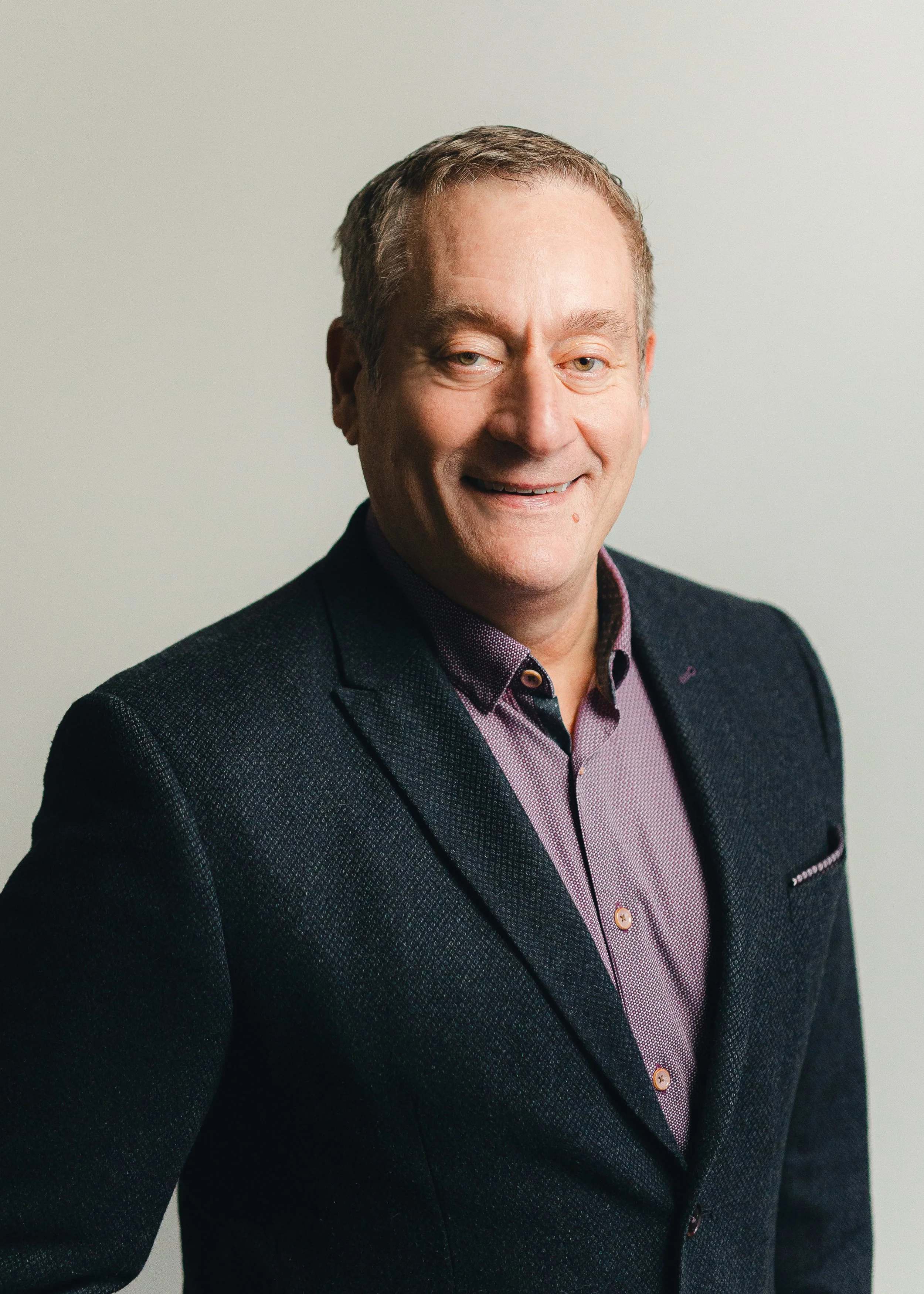 Close-up of a smiling man wearing a dark blazer and a purple patterned shirt, against a plain light background.