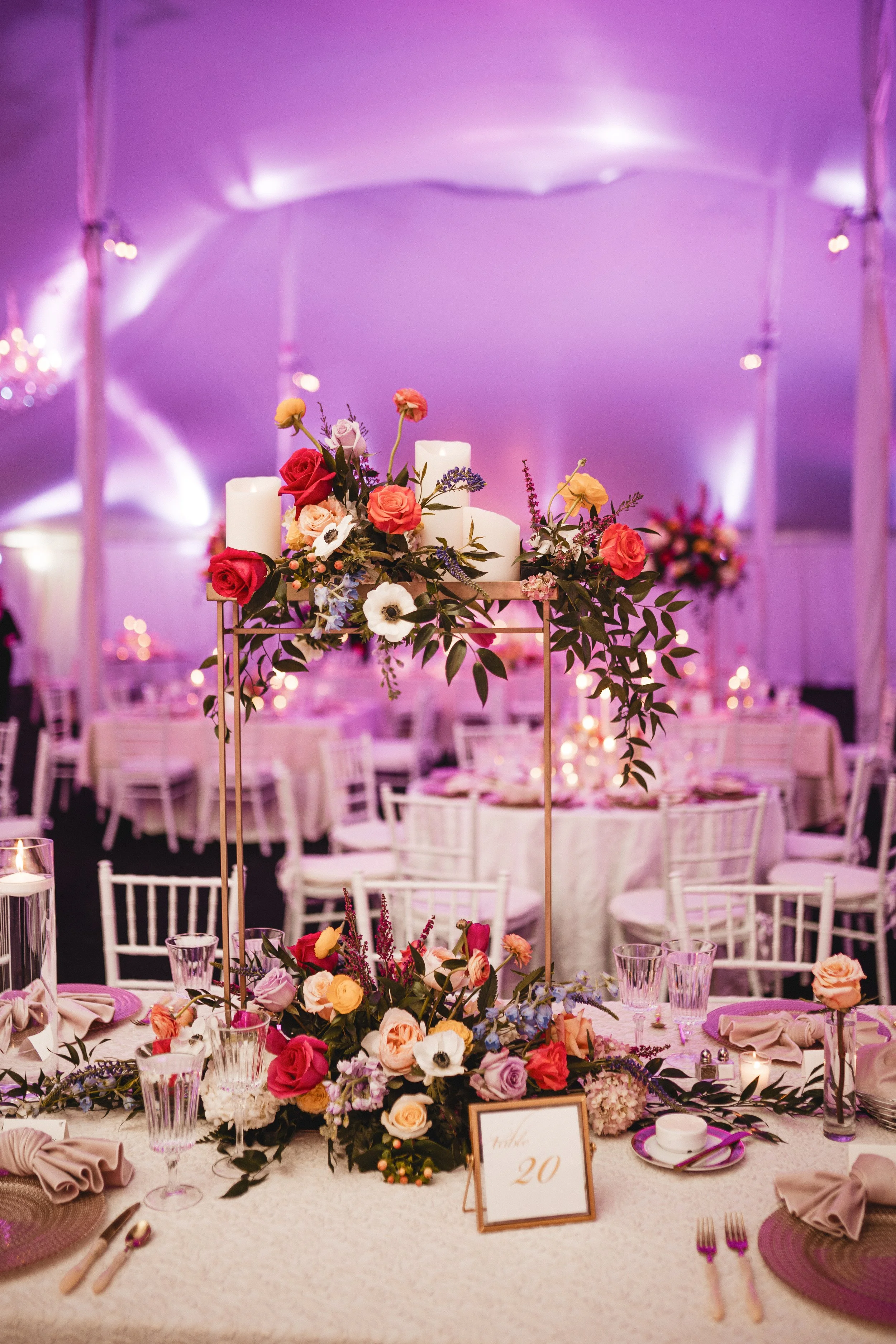 Elegant wedding reception table decorated with floral centerpiece, candles, glassware, and place settings, with a purple-lit tent ceiling in the background.