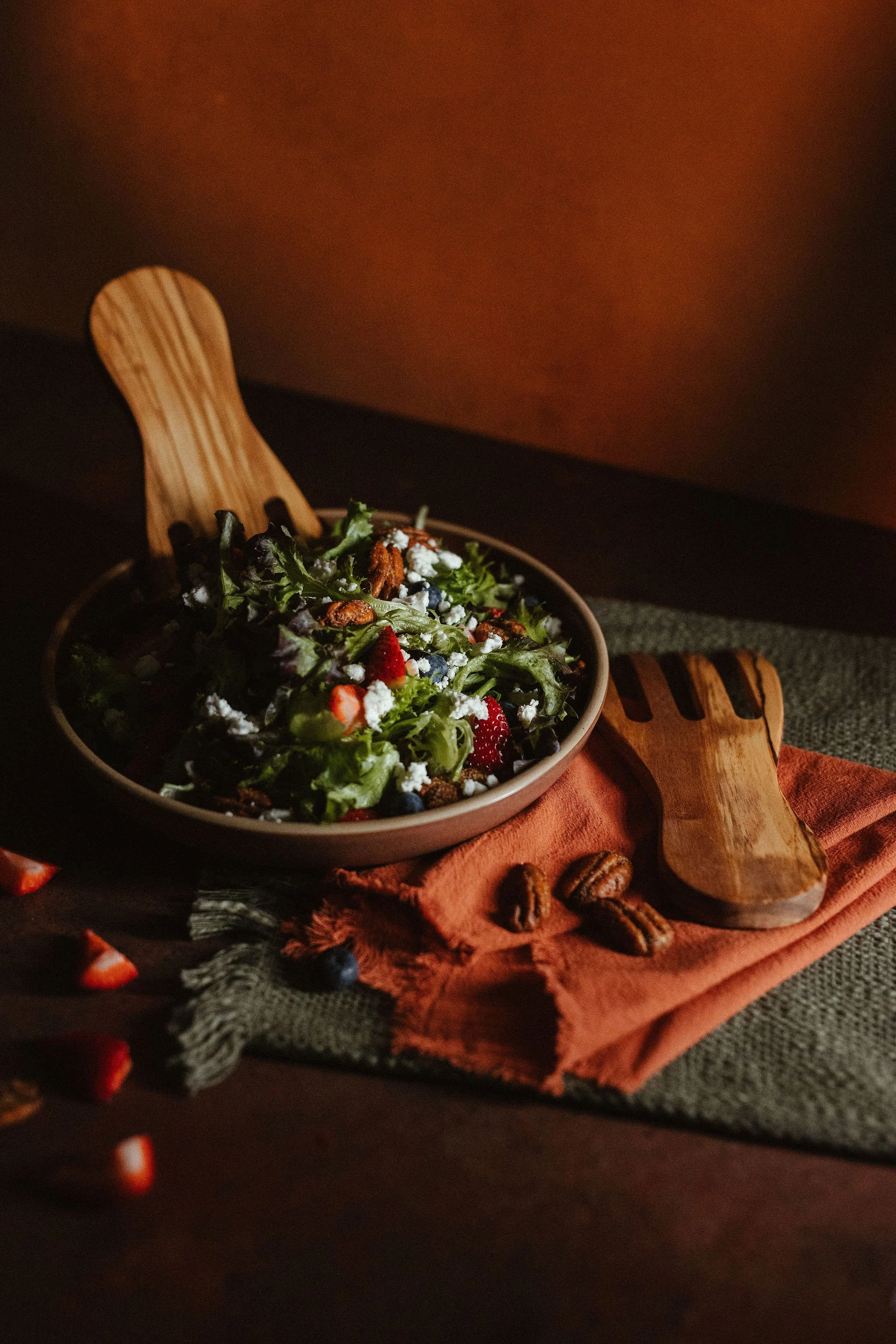 A bowl of salad containing mixed greens, strawberries, blueberries, pecans, and crumbled cheese on a dark surface with a wooden salad fork and scattered pecans.