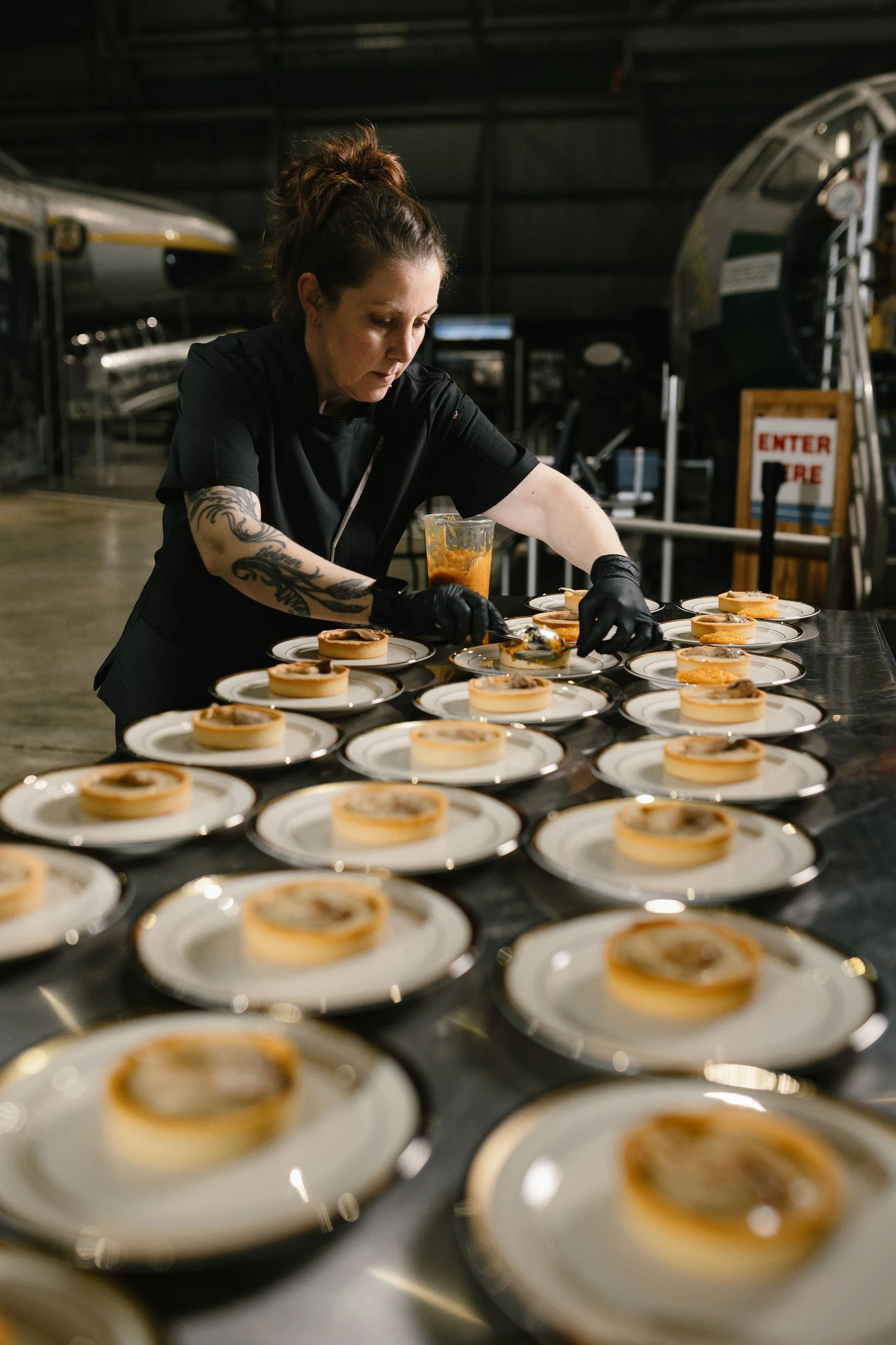 A woman wearing black gloves is preparing desserts on small white plates in an industrial kitchen or bakery setting.
