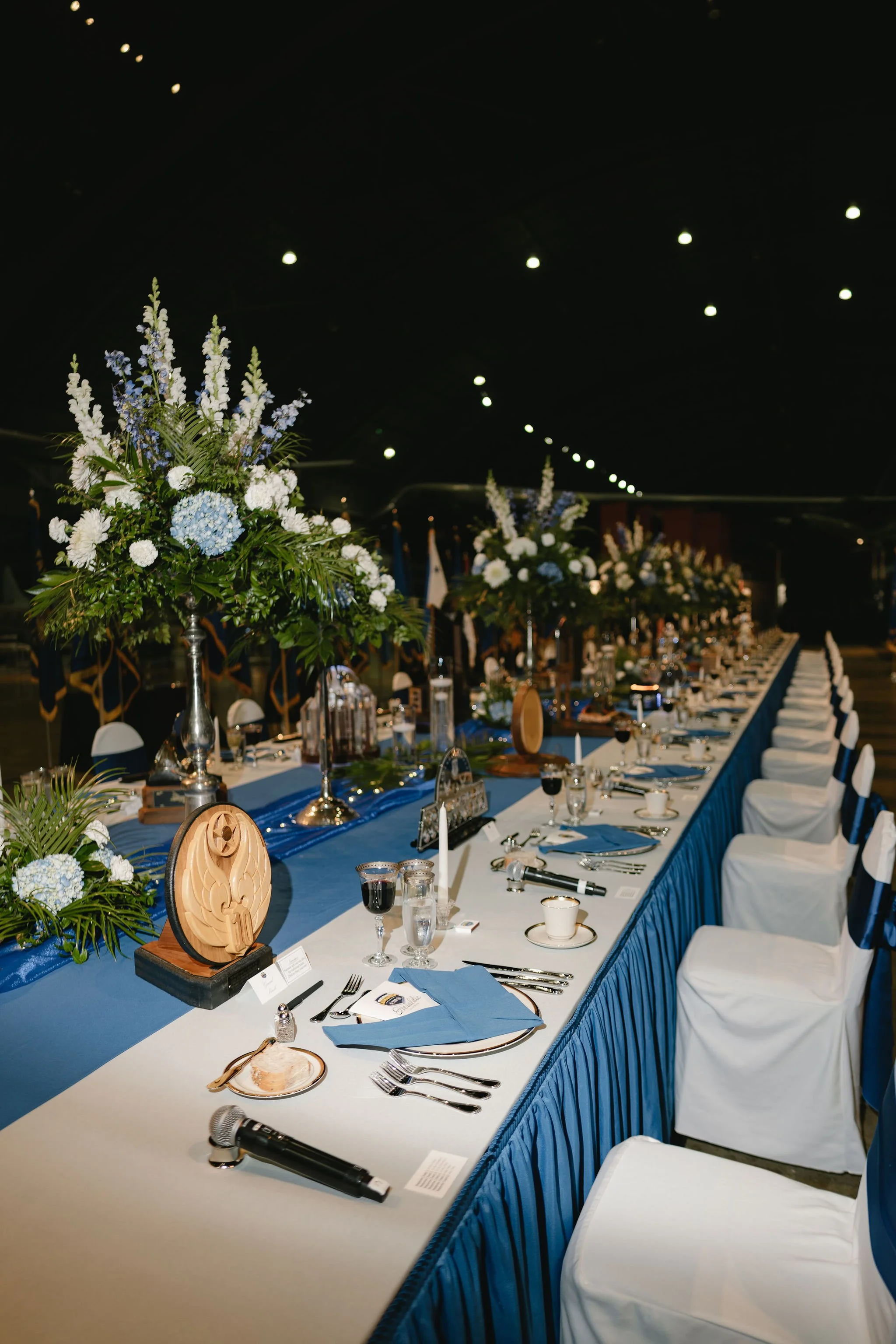Long banquet table set for a formal event, decorated with blue and white flowers, blue table runners, and place settings with glassware and silverware, in a dimly lit room with floral arrangements and decorative medals on the table.
