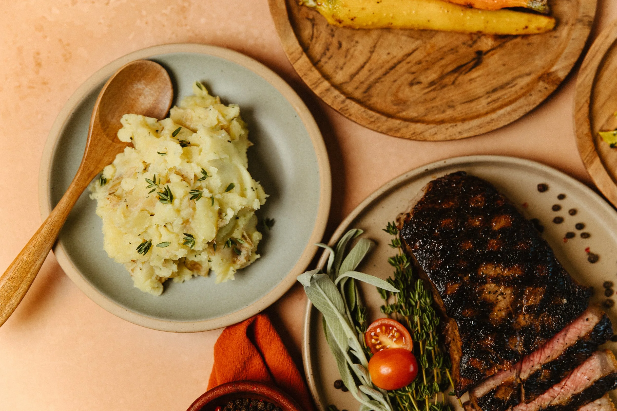 A plate of mashed potatoes garnished with herbs, a grilled steak with a dark crust, cherry tomatoes, and fresh herbs, with a wooden plate of grilled yellow squash in the background.