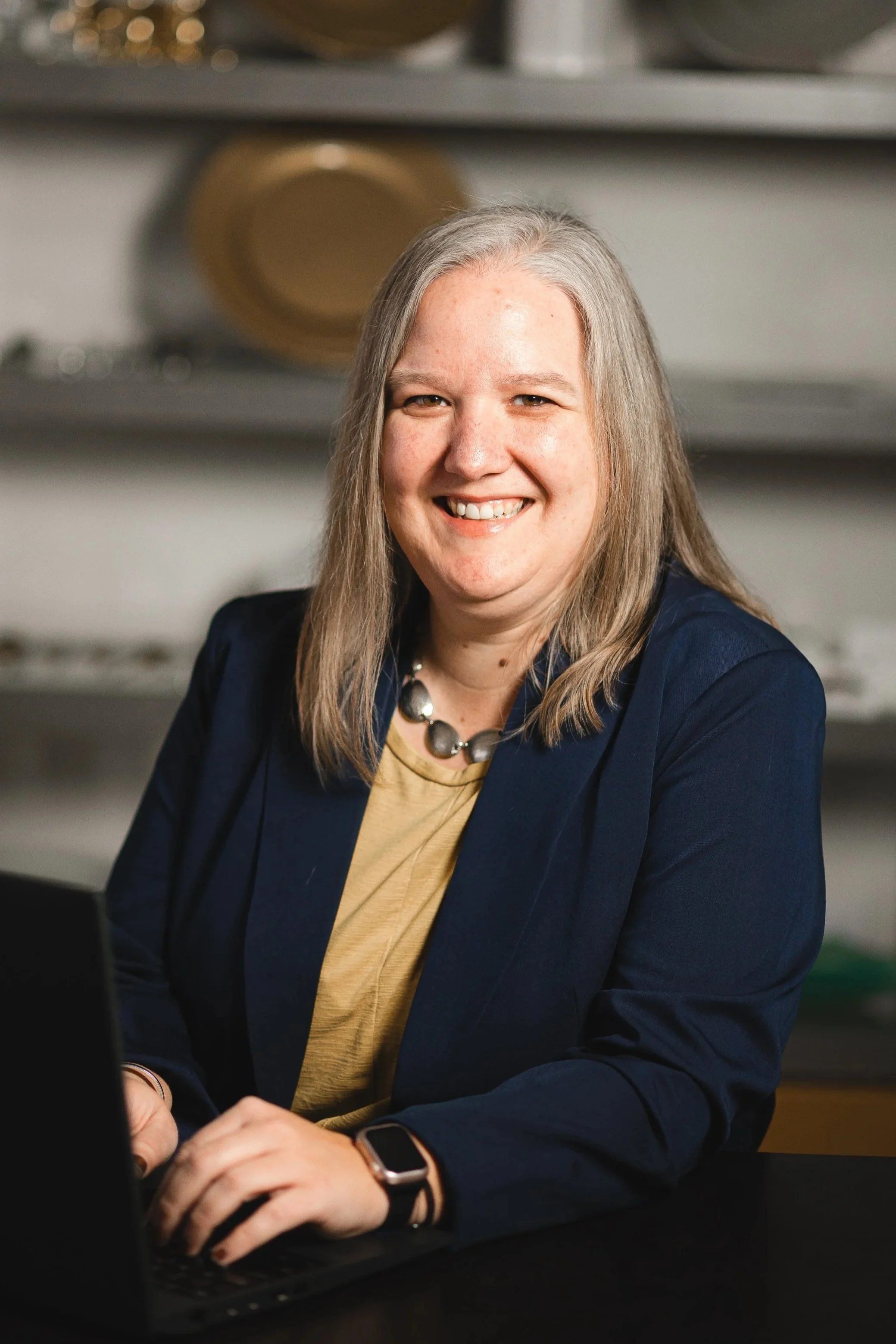 A smiling woman with long gray hair sitting at a desk with a computer in front of her in a professional setting.