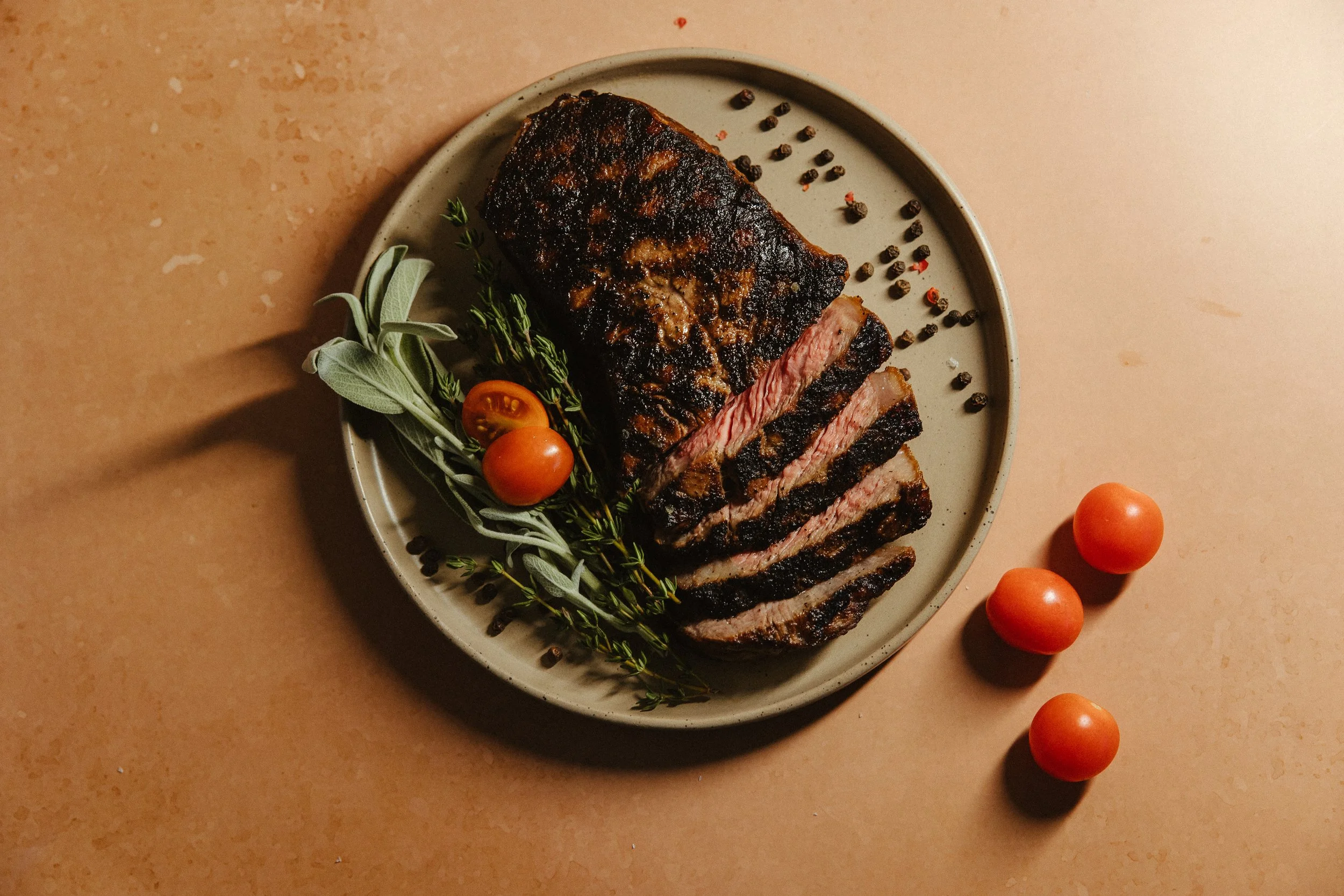 Sliced cooked steak with a charred crust on a beige plate, garnished with cherry tomatoes, sprigs of herbs, and black peppercorns on the side, on a light brown surface.