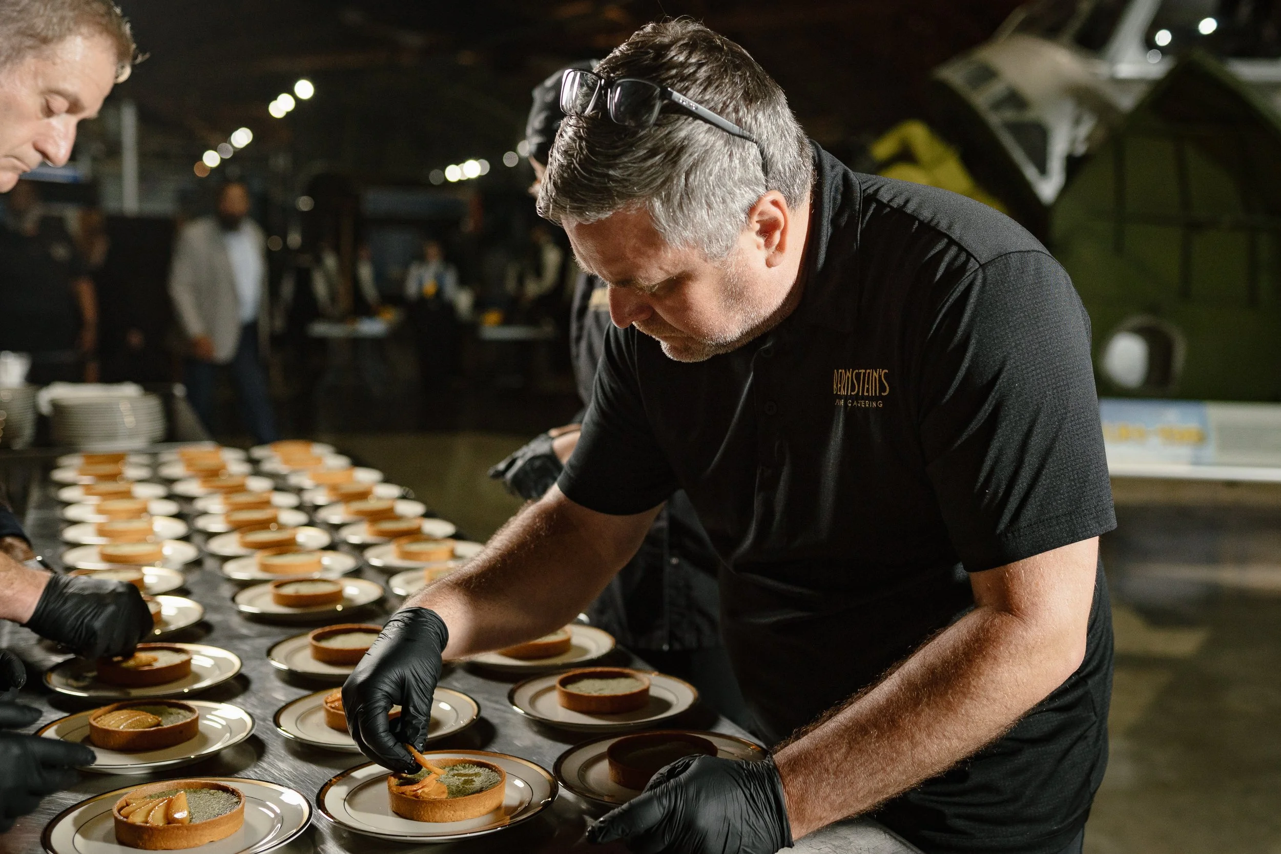 A man wearing black gloves and a black polo shirt with a logo is preparing desserts at a catering event, with several plates of tart-like desserts on a table.