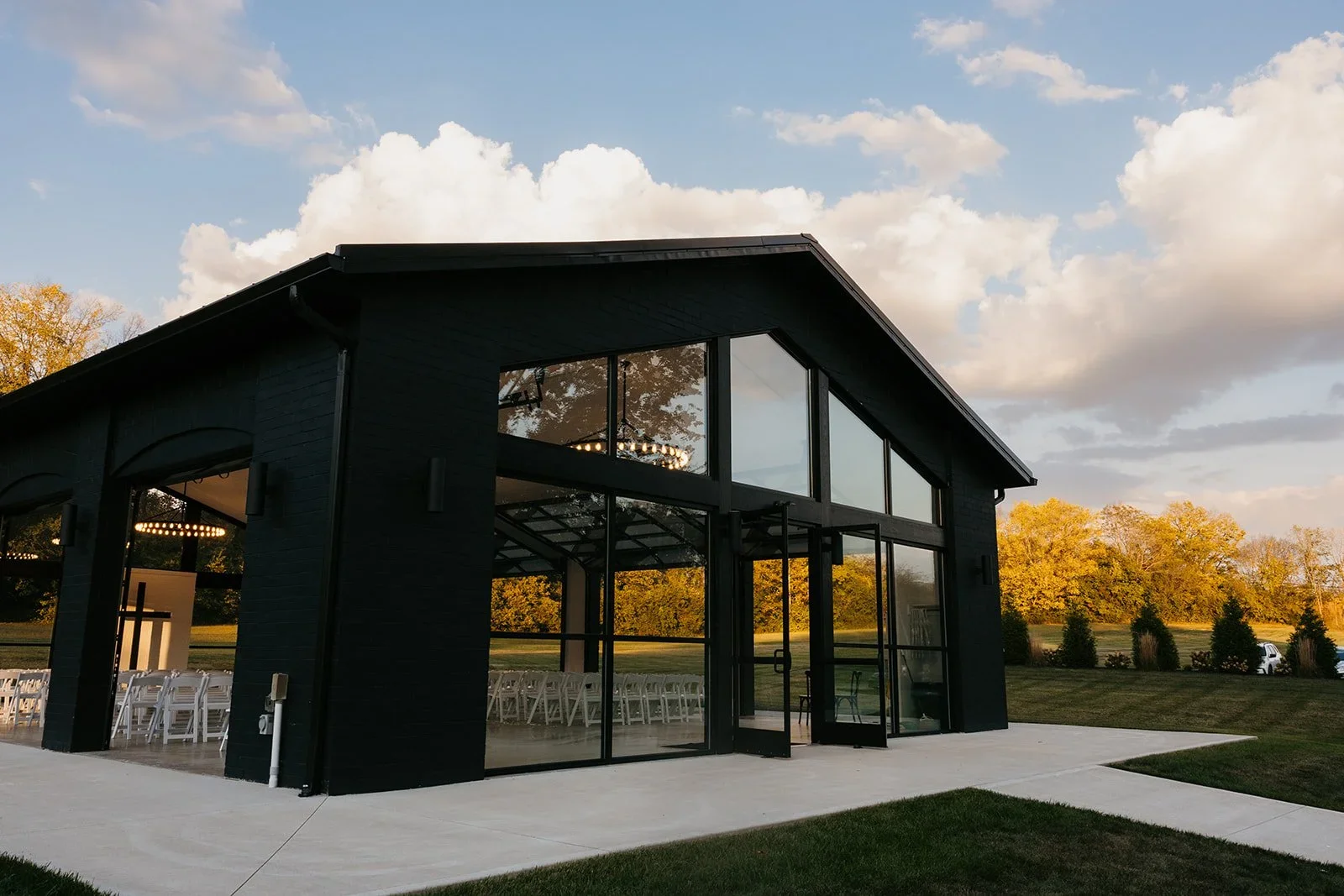 A modern black building with large glass windows and a glass entrance door, outdoor chairs arranged inside, surrounded by green grass and trees, under a partly cloudy sky.
