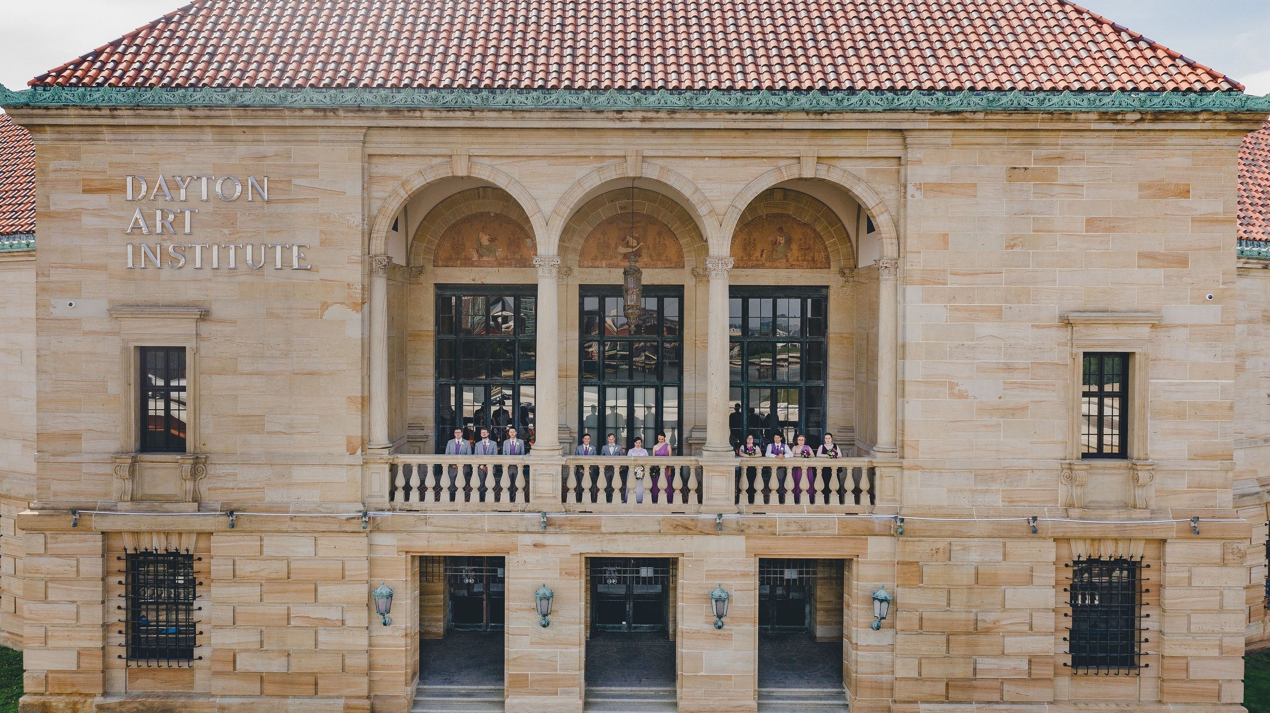 View of the Dayton Art Institute building with a group of people dressed in academic robes standing on the balcony, in front of three arched windows with murals, beige stone exterior, and a red tile roof.