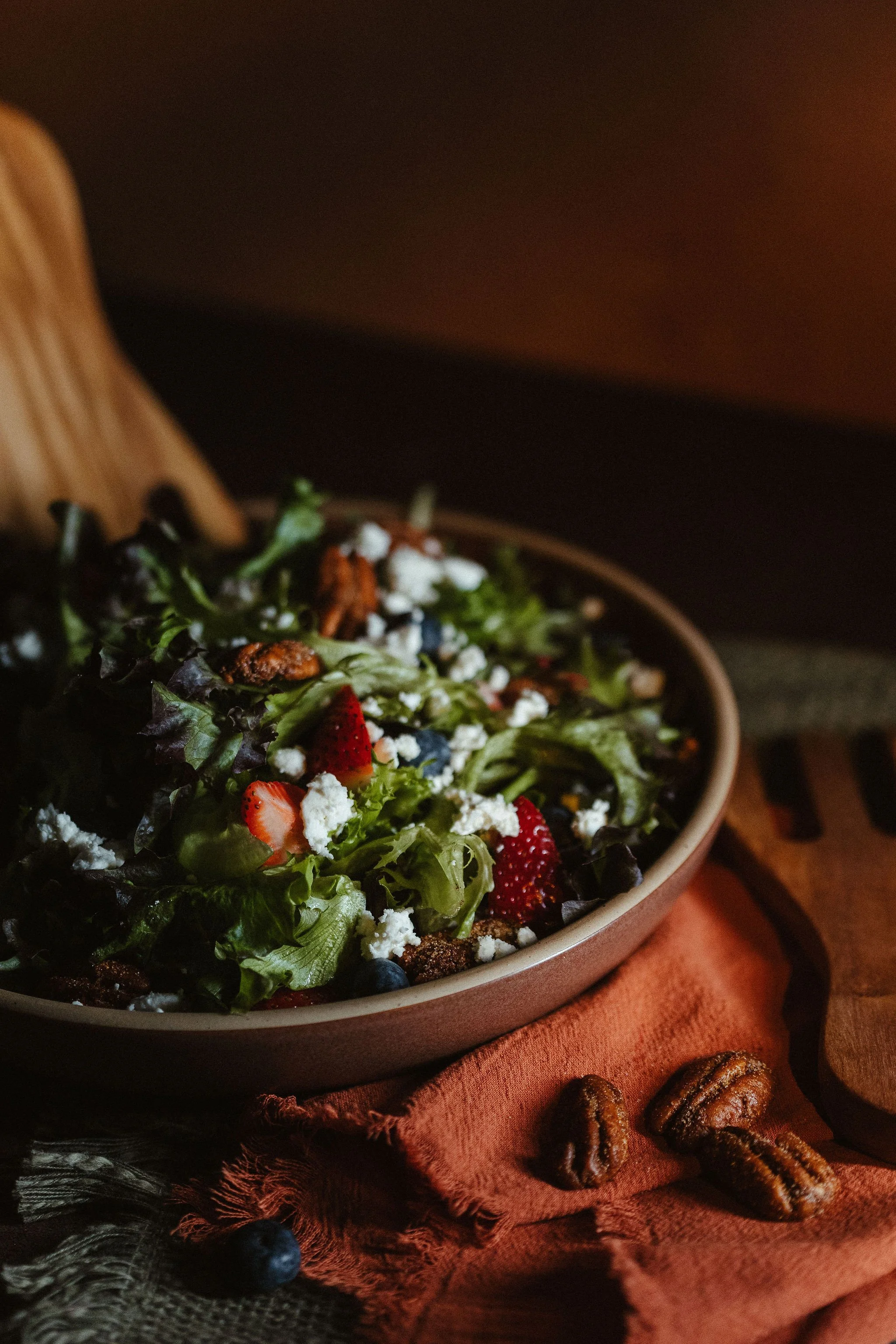 A close-up of a salad in a bowl with various greens, strawberries, blueberries, feta cheese, and nuts on a rustic table with pecans, a fork, and a cloth napkin.