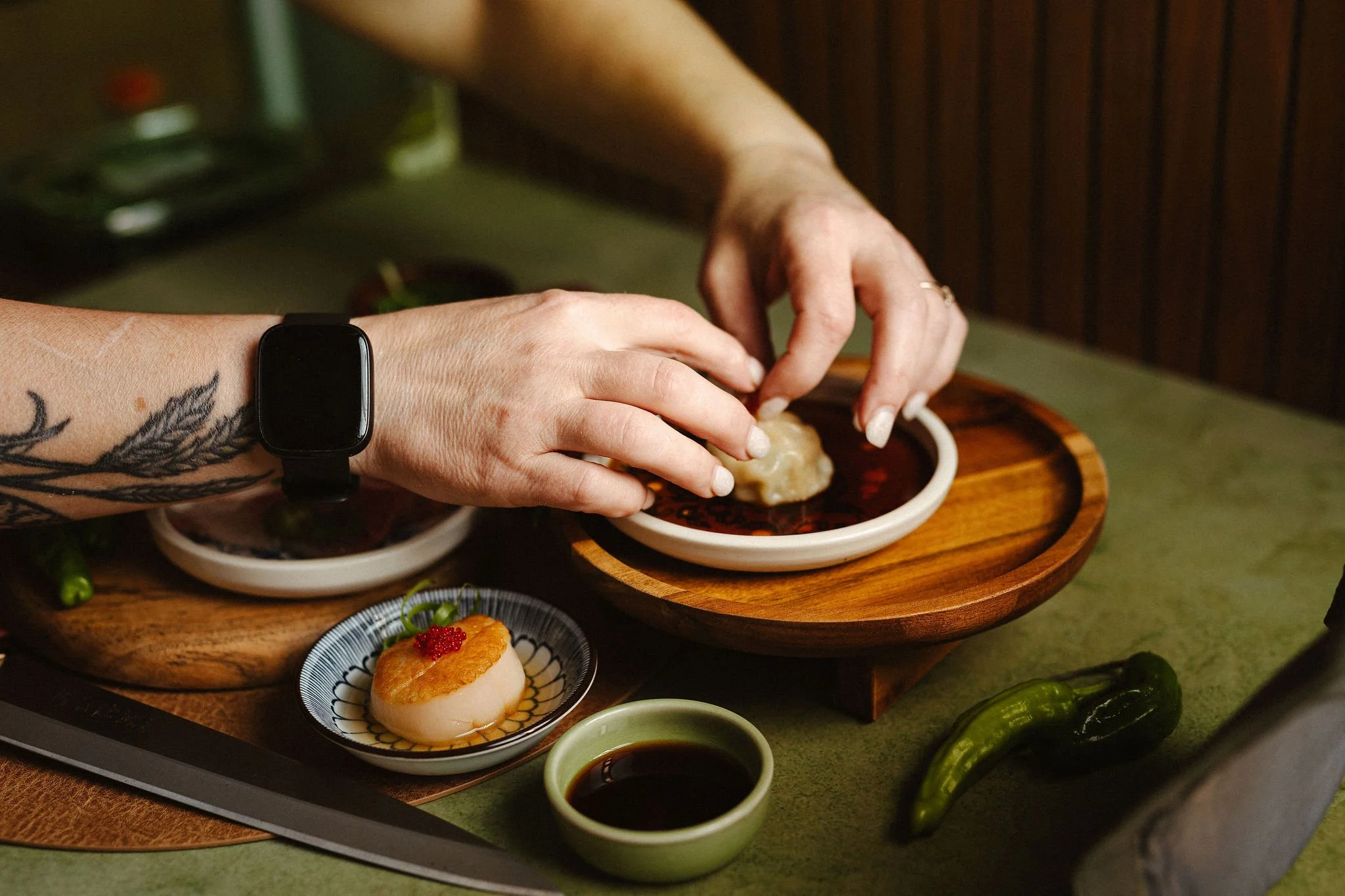 Close-up of hands preparing sushi with soy sauce and wasabi on a wooden serving tray, with a sushi roll, dipping bowls, and a dessert in the background.