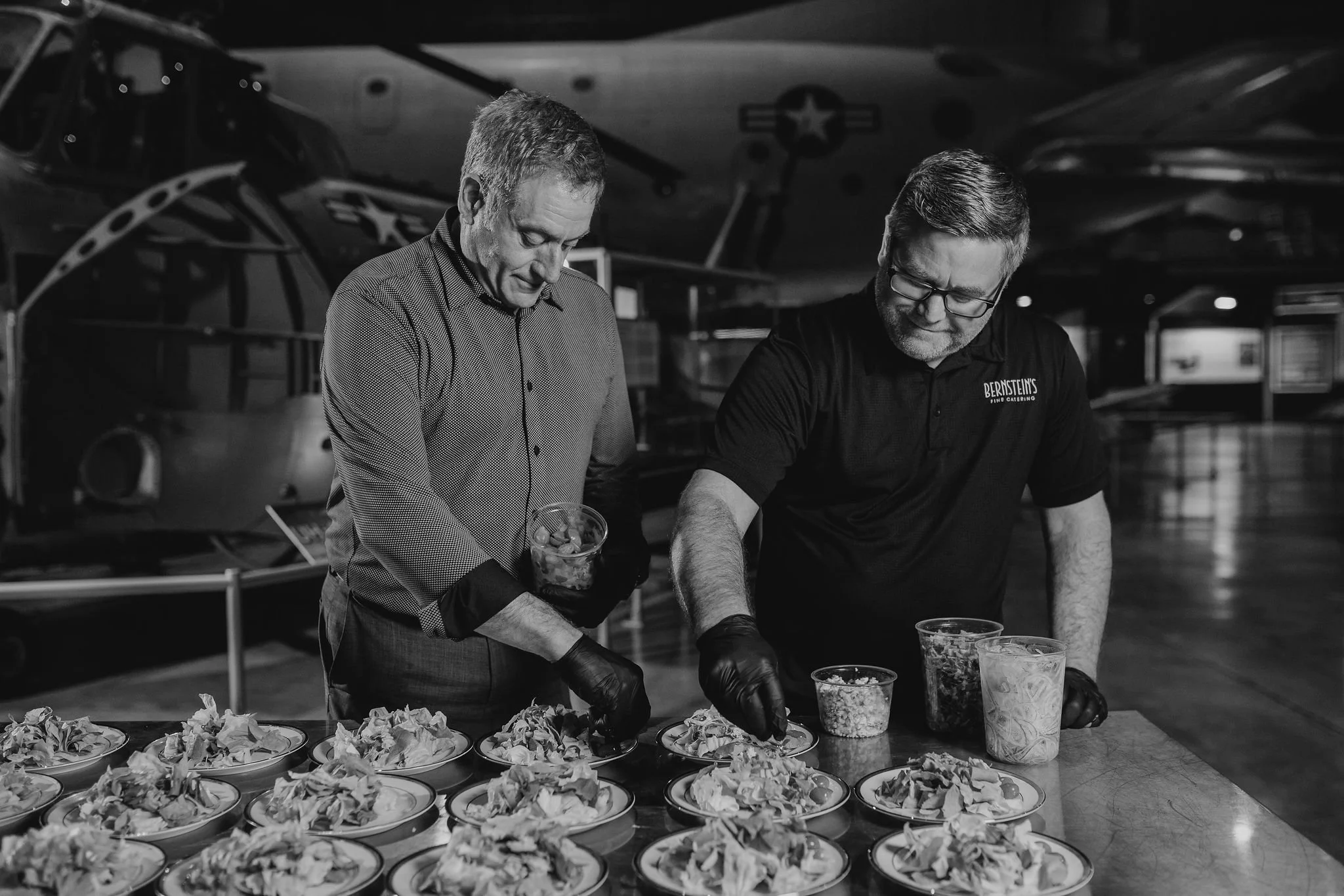 Two men preparing food at a table with various dishes, in an aviation museum with airplanes in the background.