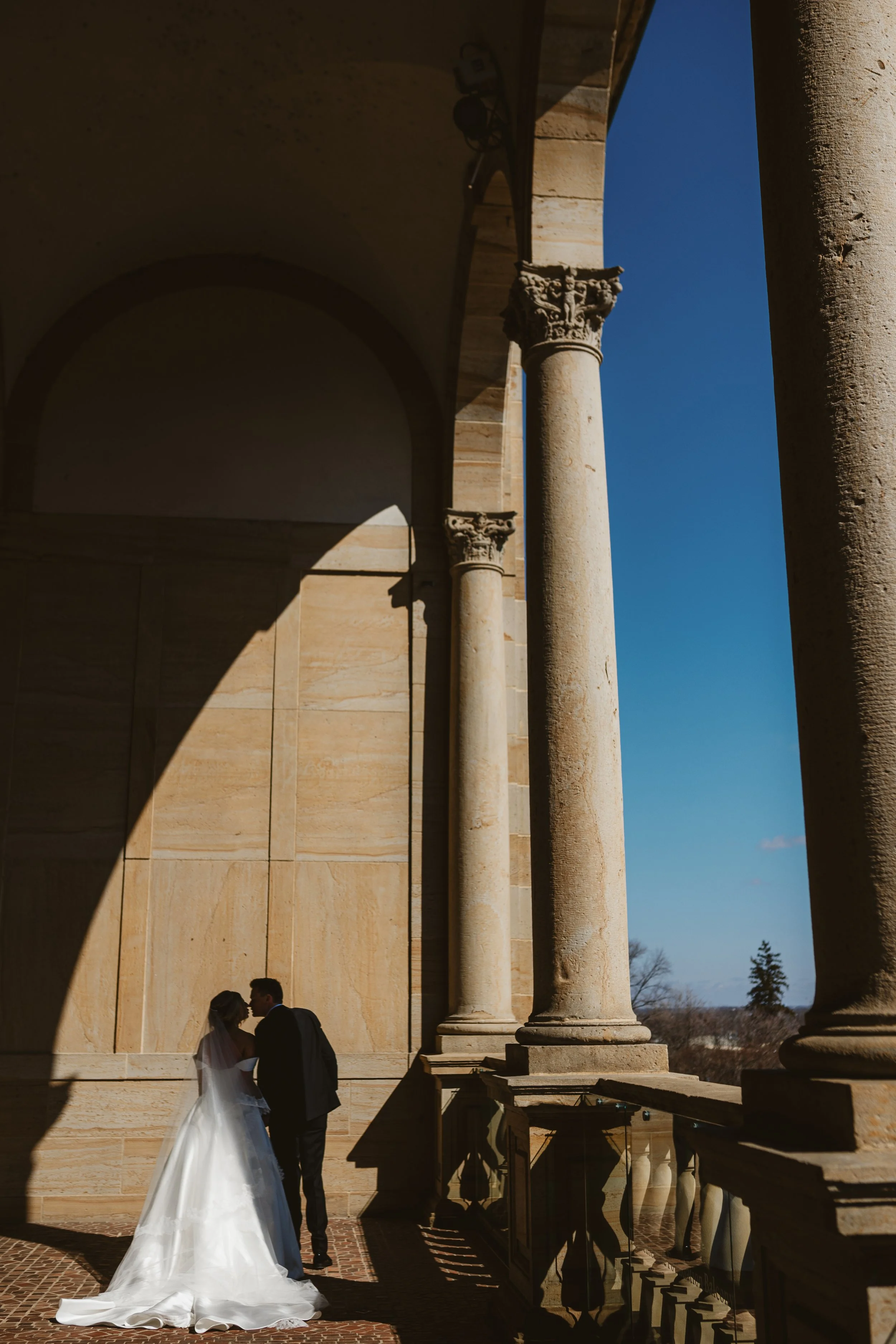 A bride and groom stand close together in front of ancient stone columns and columns casting shadows on a building, under a clear blue sky, during a wedding ceremony.