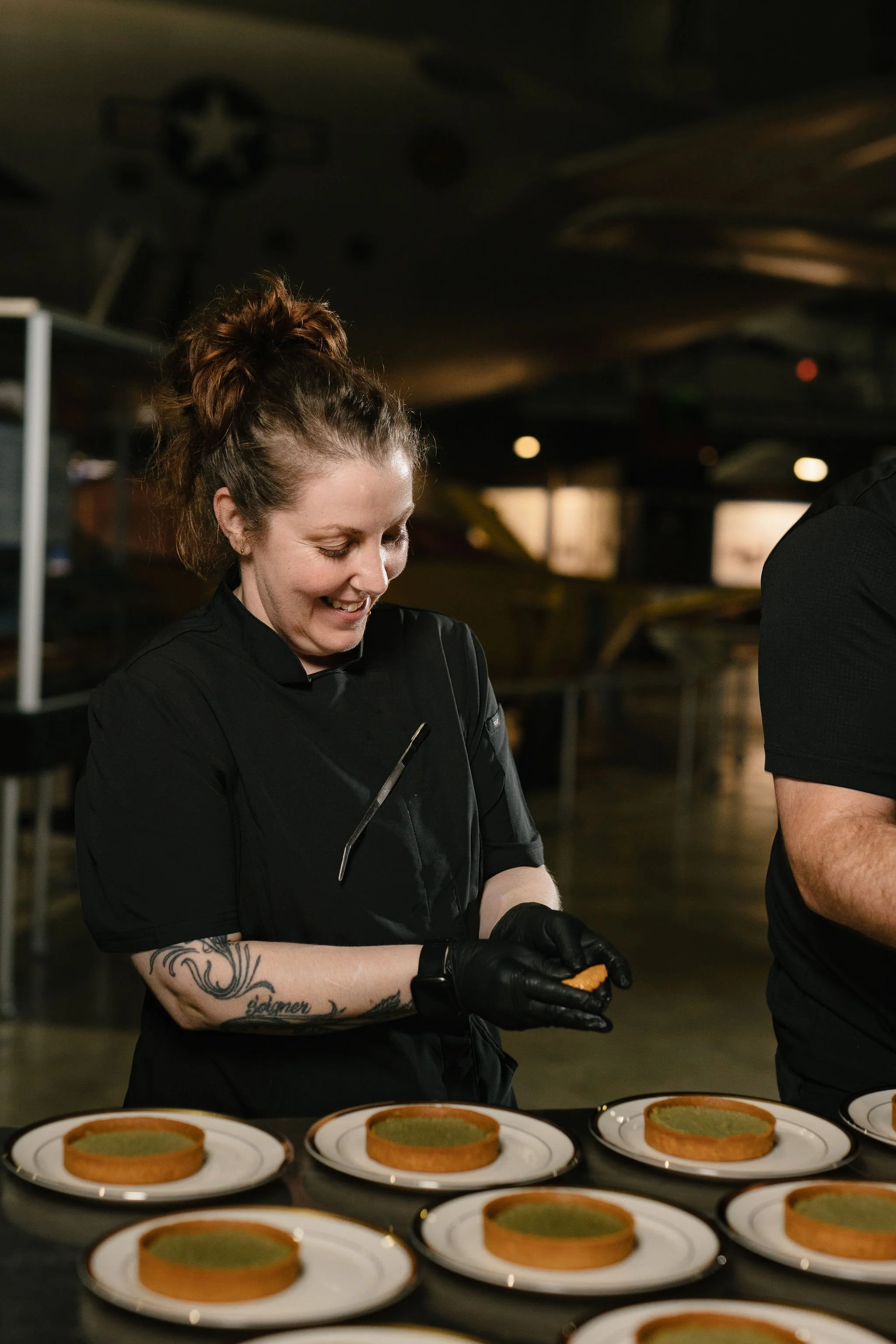 A woman wearing a black uniform and black gloves stands in a kitchen, smiling and preparing food with several plates of green tart on a table in front of her.
