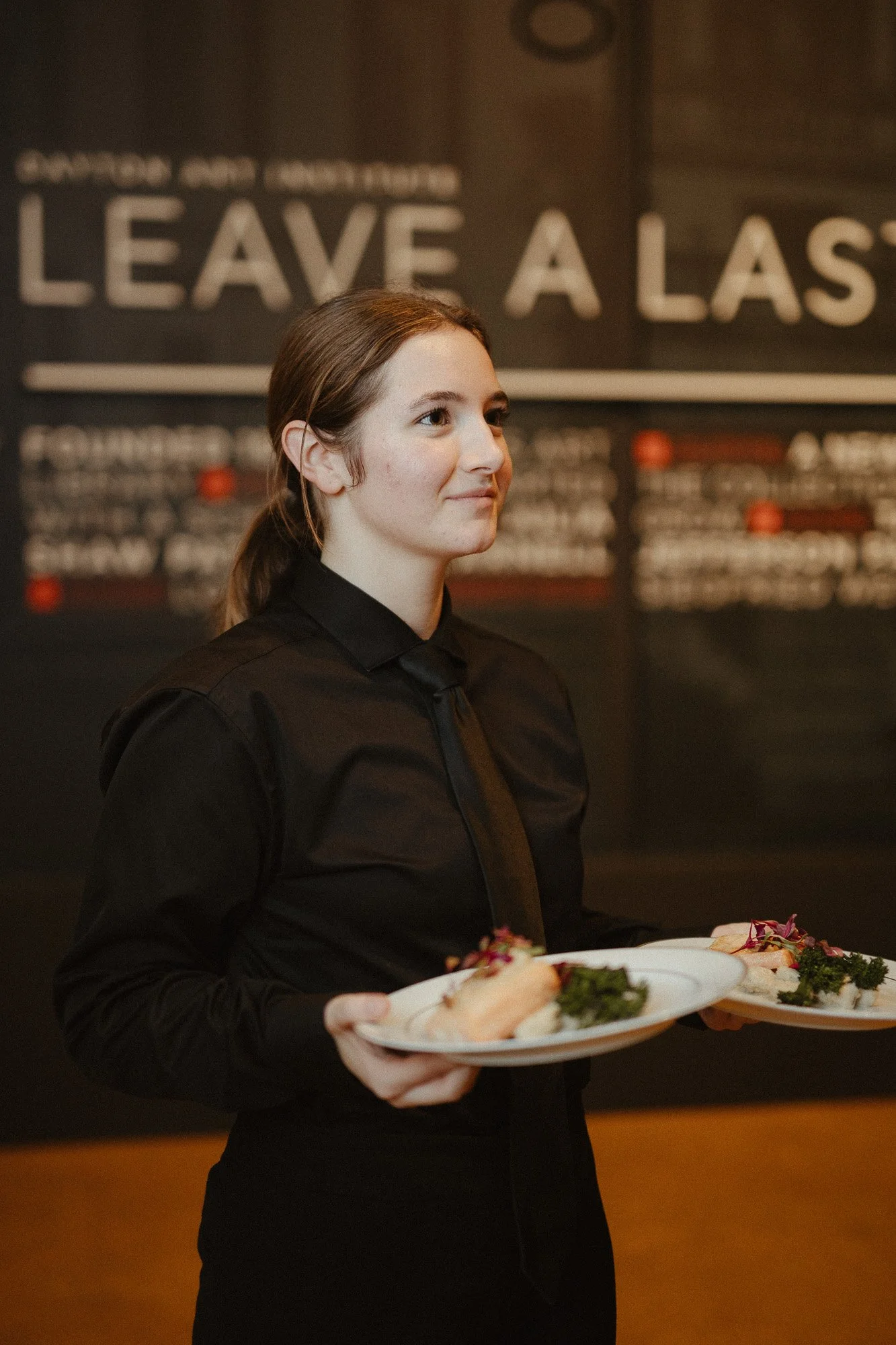 A young woman with brown hair in a ponytail, wearing a black uniform, holding a plate with food, standing indoors with a black wall featuring white text behind her.