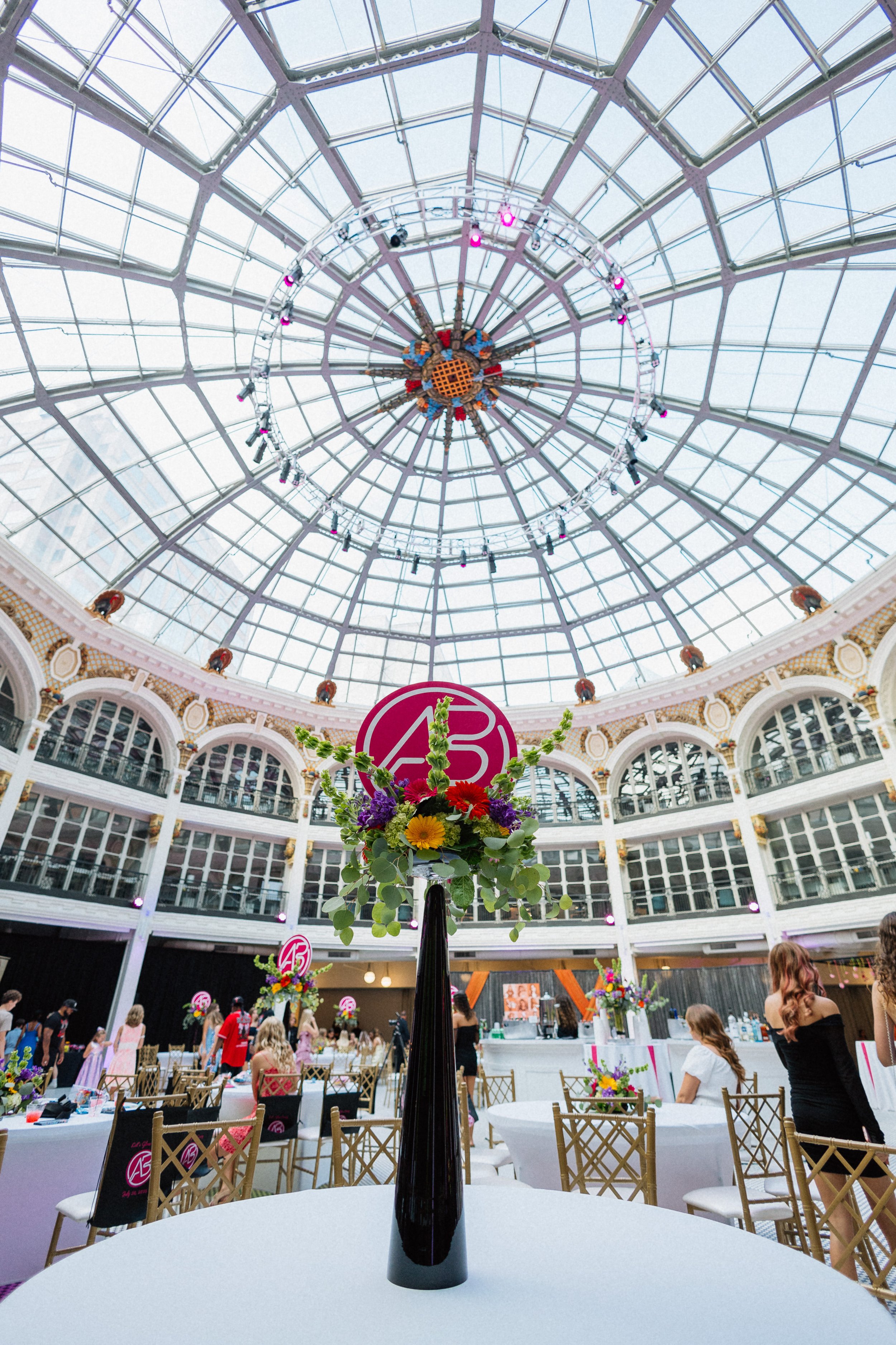 A tall black vase with a bouquet of colorful flowers on a white table in the center of a large, bright ballroom with a glass domed ceiling. People are gathered around, and pink lighting is illuminating parts of the ceiling.