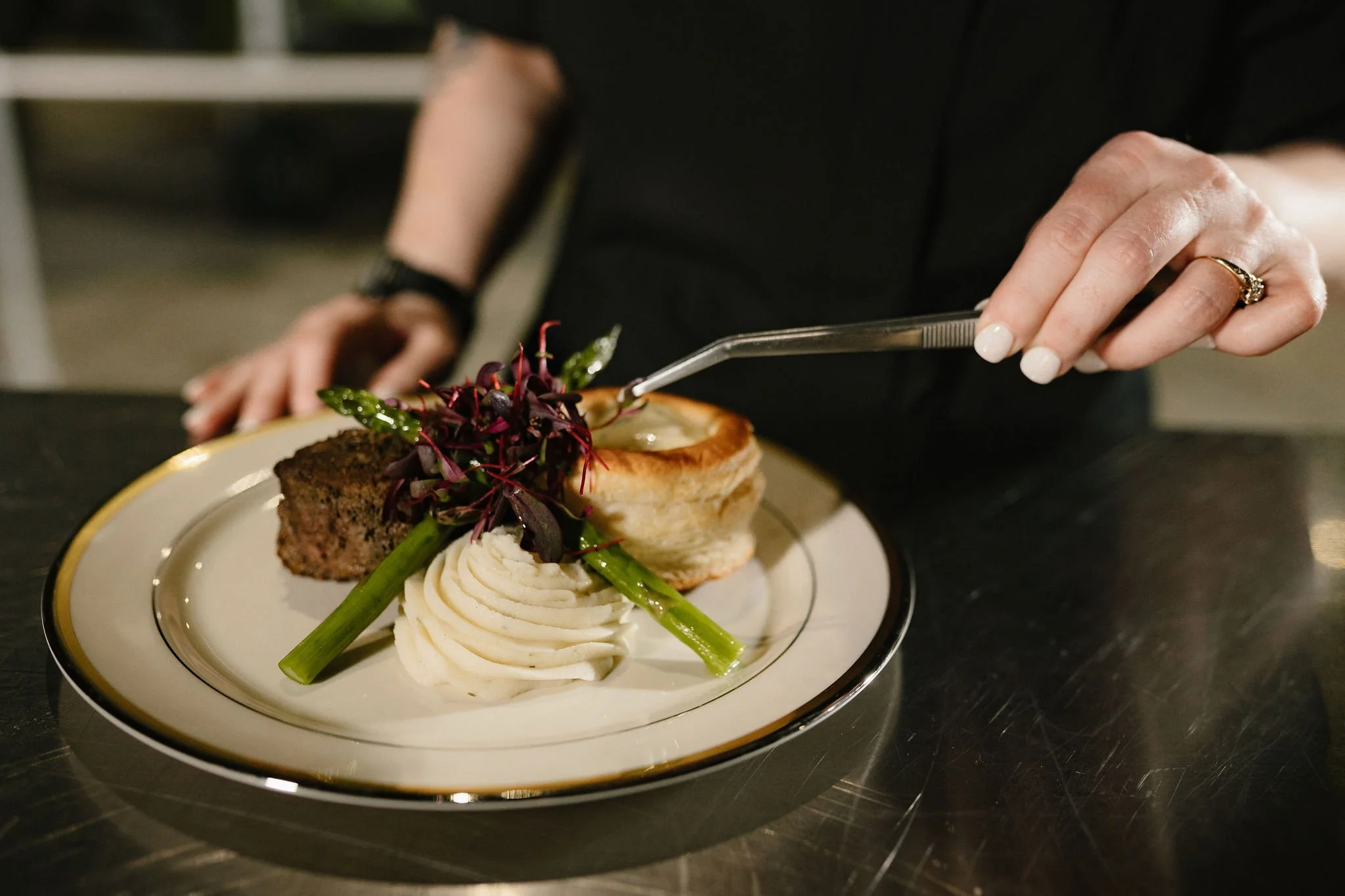 A plate of food with mashed potatoes, green beans, a slice of meatloaf, and a small salad, being served with a fork by a person wearing a black outfit and a ring.
