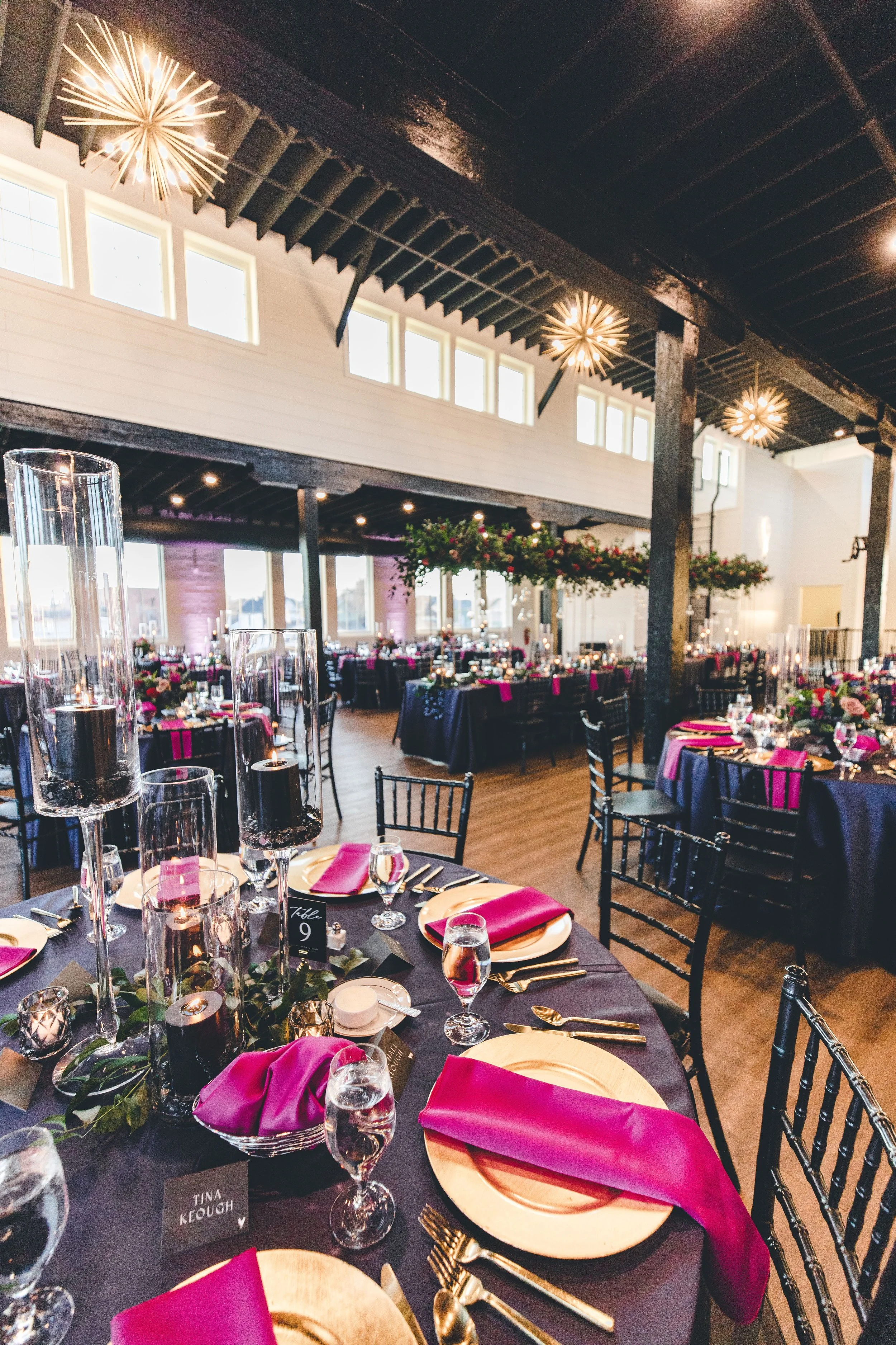 Interior of an event hall decorated for a celebration with round tables, pink napkins, candles, and floral arrangements. Modern chandeliers hang from a dark ceiling, and natural light streams in through large windows.