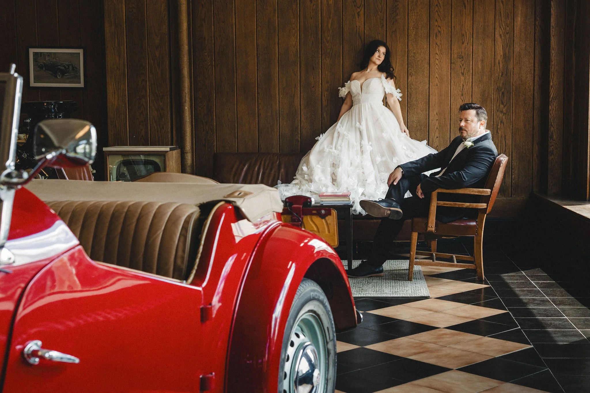 A woman in a wedding dress standing next to a seated man in a dark suit inside a wooden-paneled room with vintage decor, and a red vintage car in the foreground.