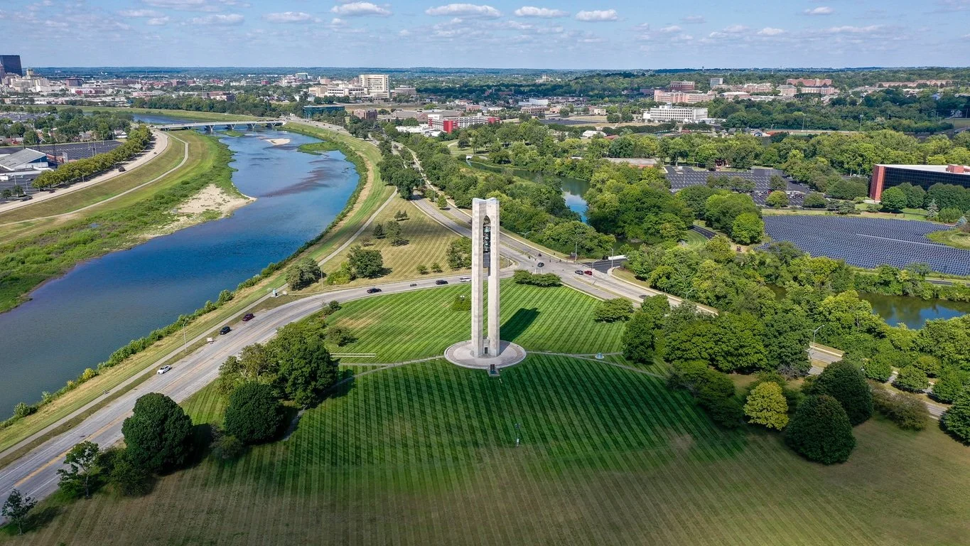 Aerial view of a city park with a tall monument, a curved road, a river, green trees, and surrounding cityscape.