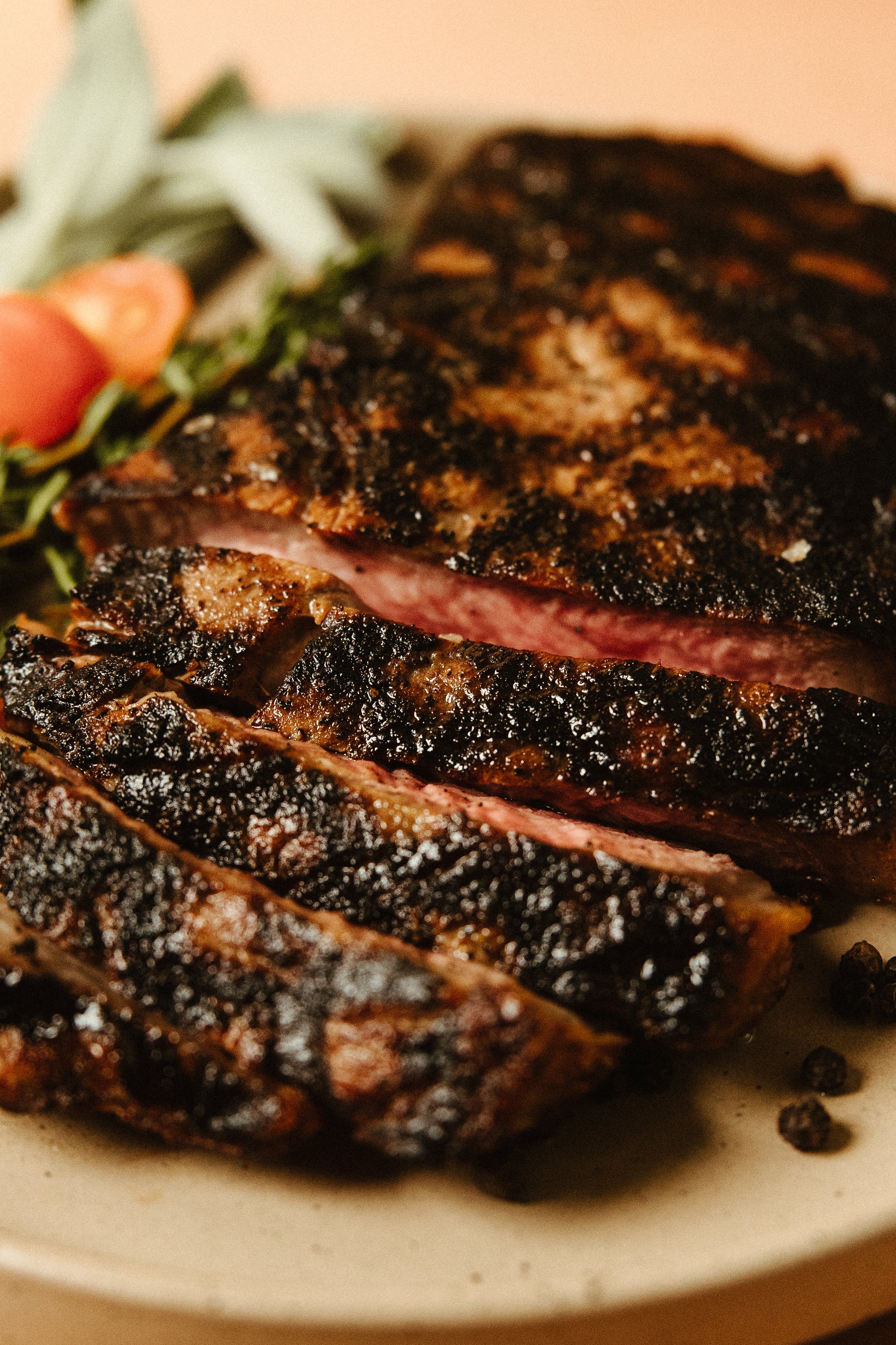 Close-up of a steak with a charred, crispy exterior and a hint of pink in the interior, on a white plate with some black peppercorns.