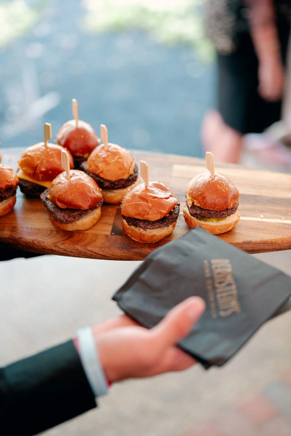 A wooden serving board with mini burger sliders topped with small pick sticks, set against a blurred background. A person in a black jacket is holding a dark napkin in their hand.